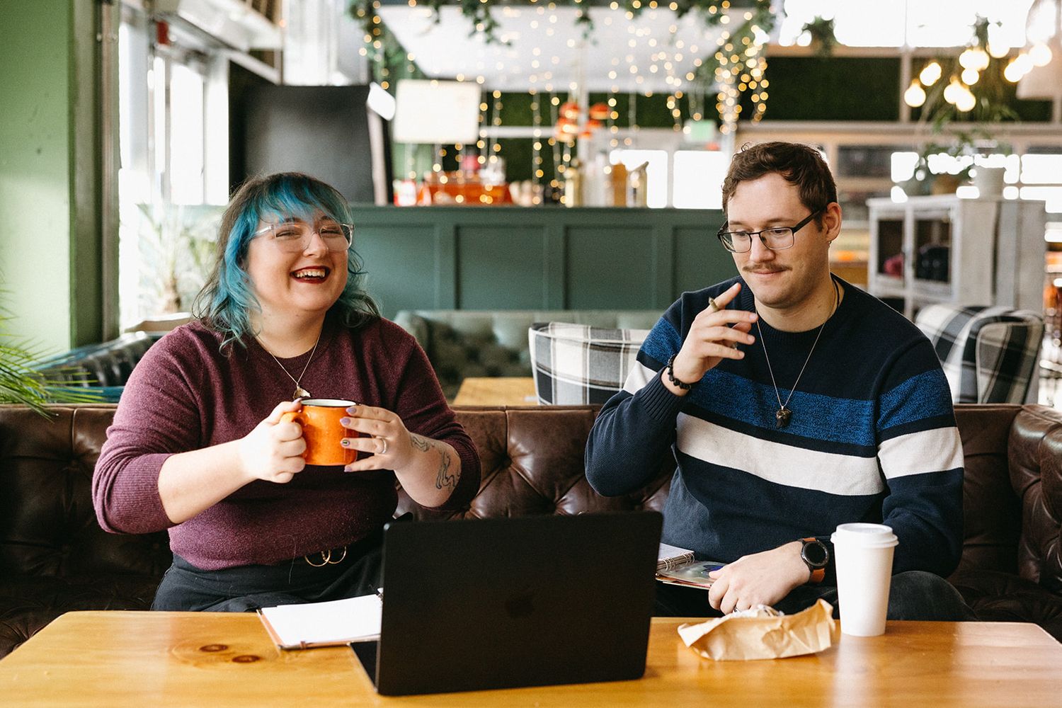 Two people sit at a cafe table with coffee cups and a laptop, having a casual meeting in a bright modern space.