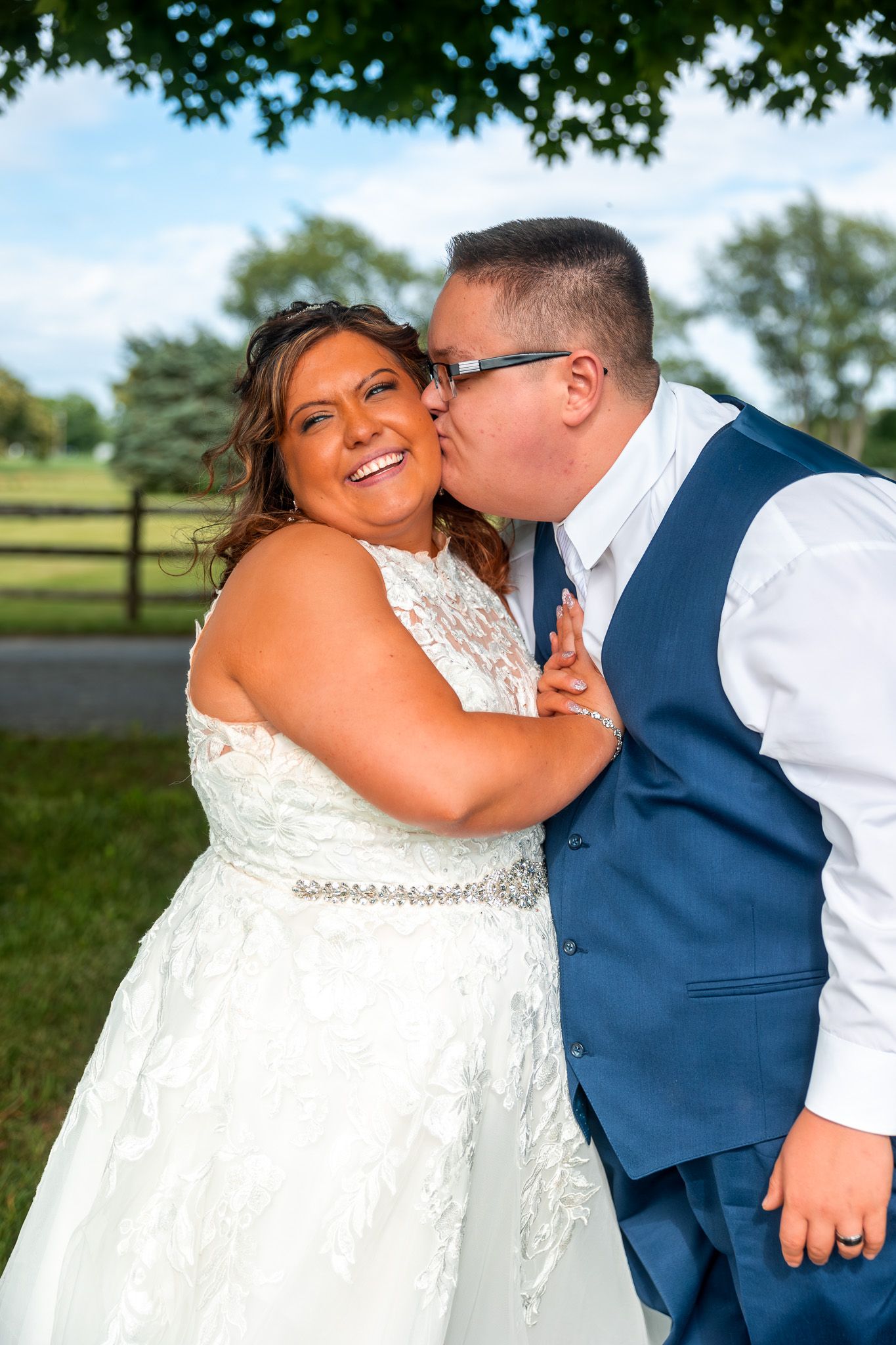 A joyful newlywed couple shares a tender moment outdoors on their wedding day with trees and fencing in the background.