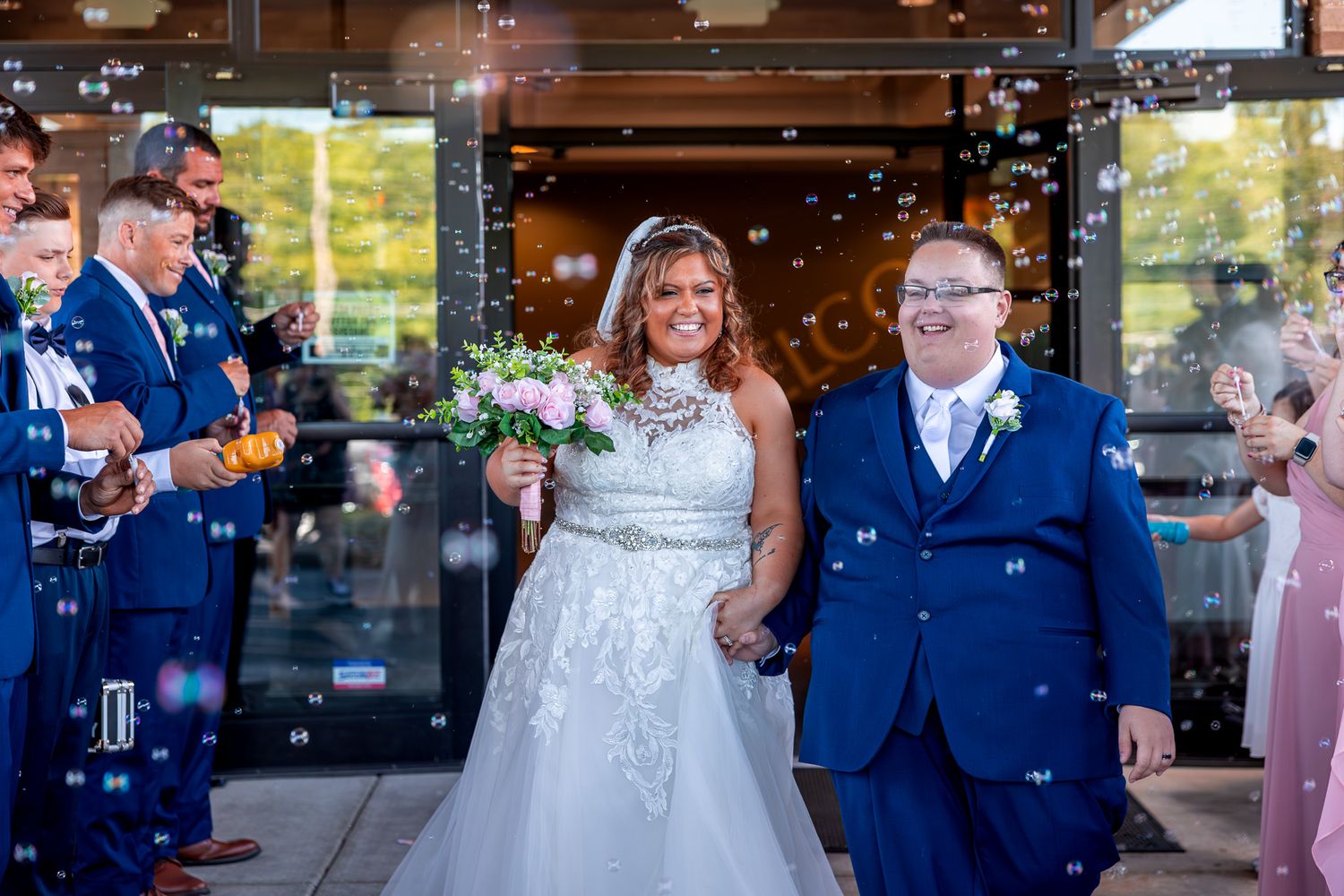 A joyful wedding couple walks down the aisle under falling bubbles with guests in blue attire and pink dresses.
