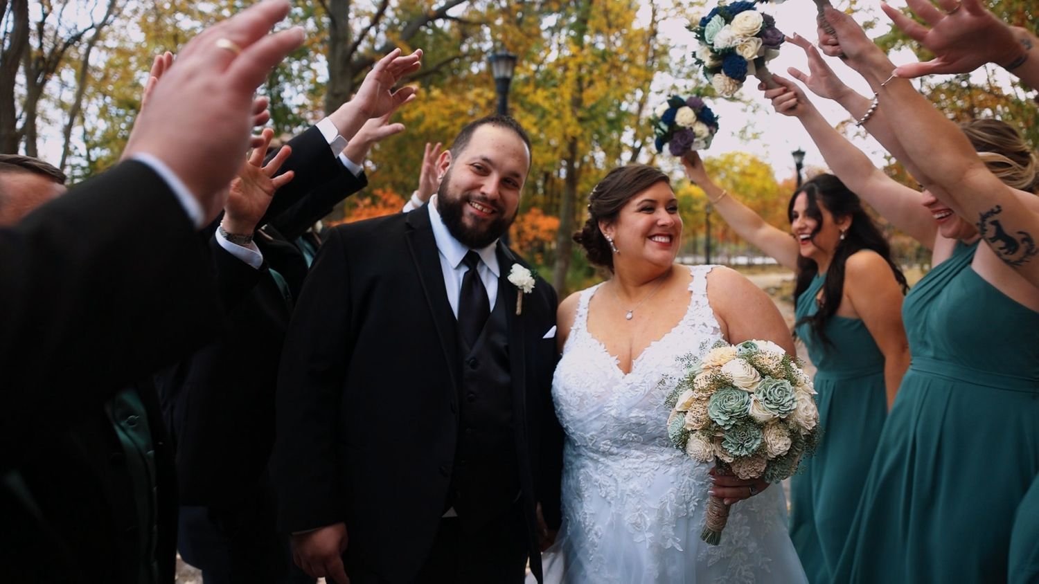 Happy wedding couple smiles while surrounded by celebrating guests in teal dresses during an autumn outdoor celebration.