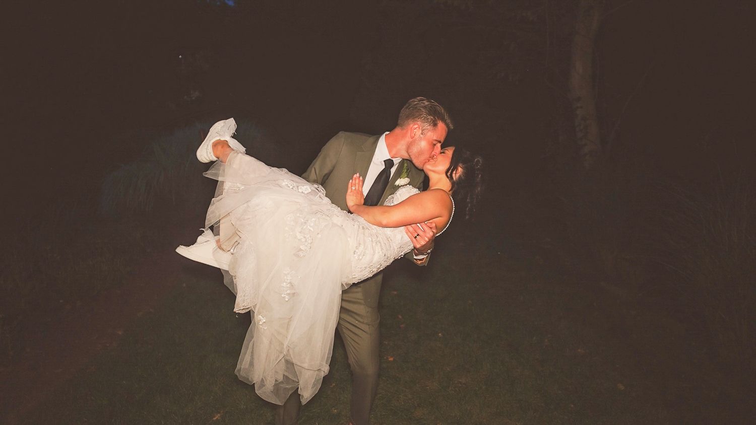 A couple in wedding attire embraces in a romantic dip pose outdoors at night during their celebration.