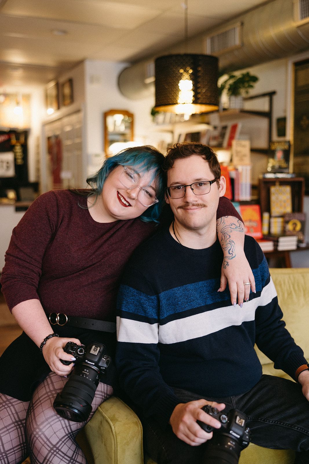 Two photographers sit together on a couch holding professional cameras in what appears to be a cozy indoor setting.