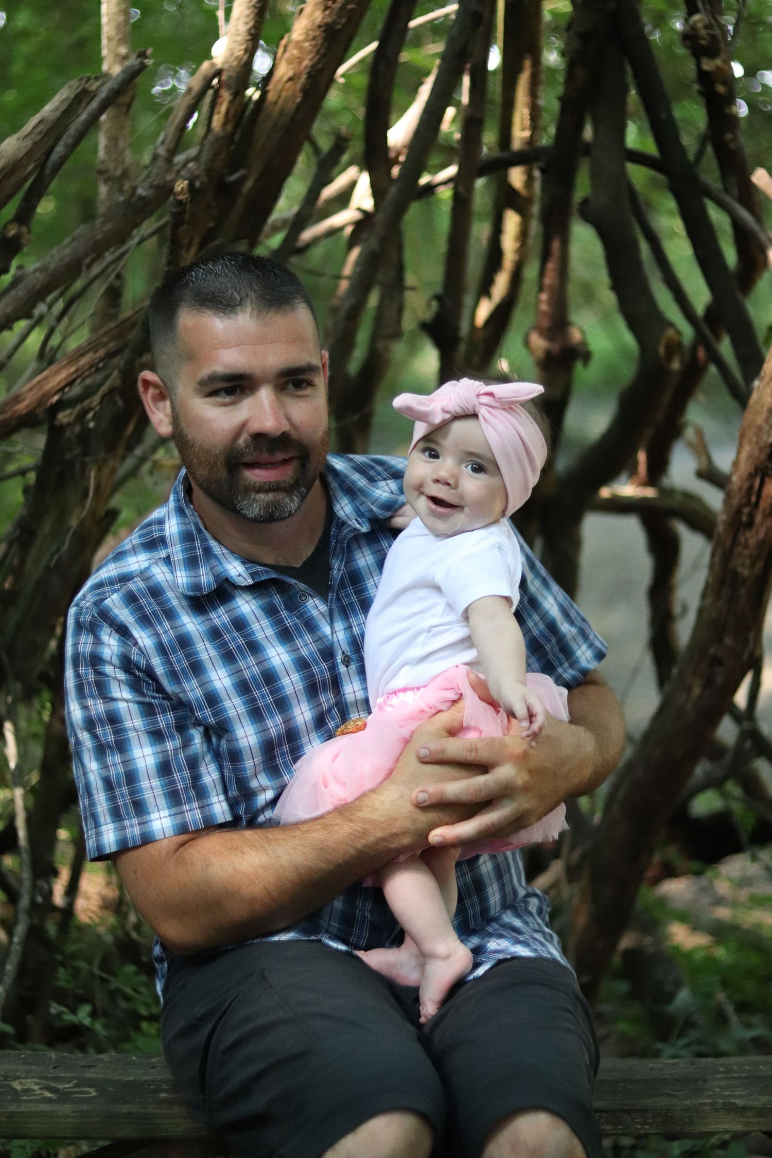 A smiling baby wearing pink pants and bow headband sits with an adult in plaid shirt under a natural wood archway outdoors.