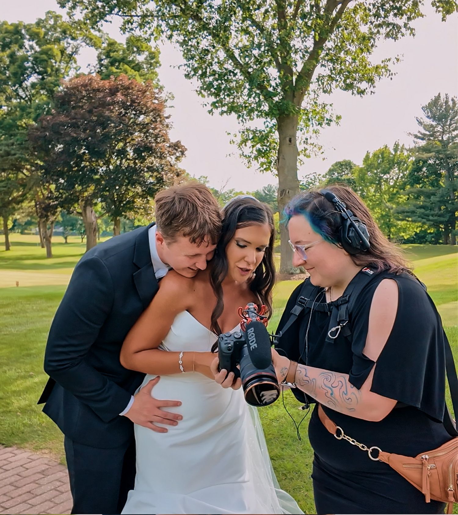A photographer shows a couple their photos on a camera display while standing on a brick pathway in a park setting.
