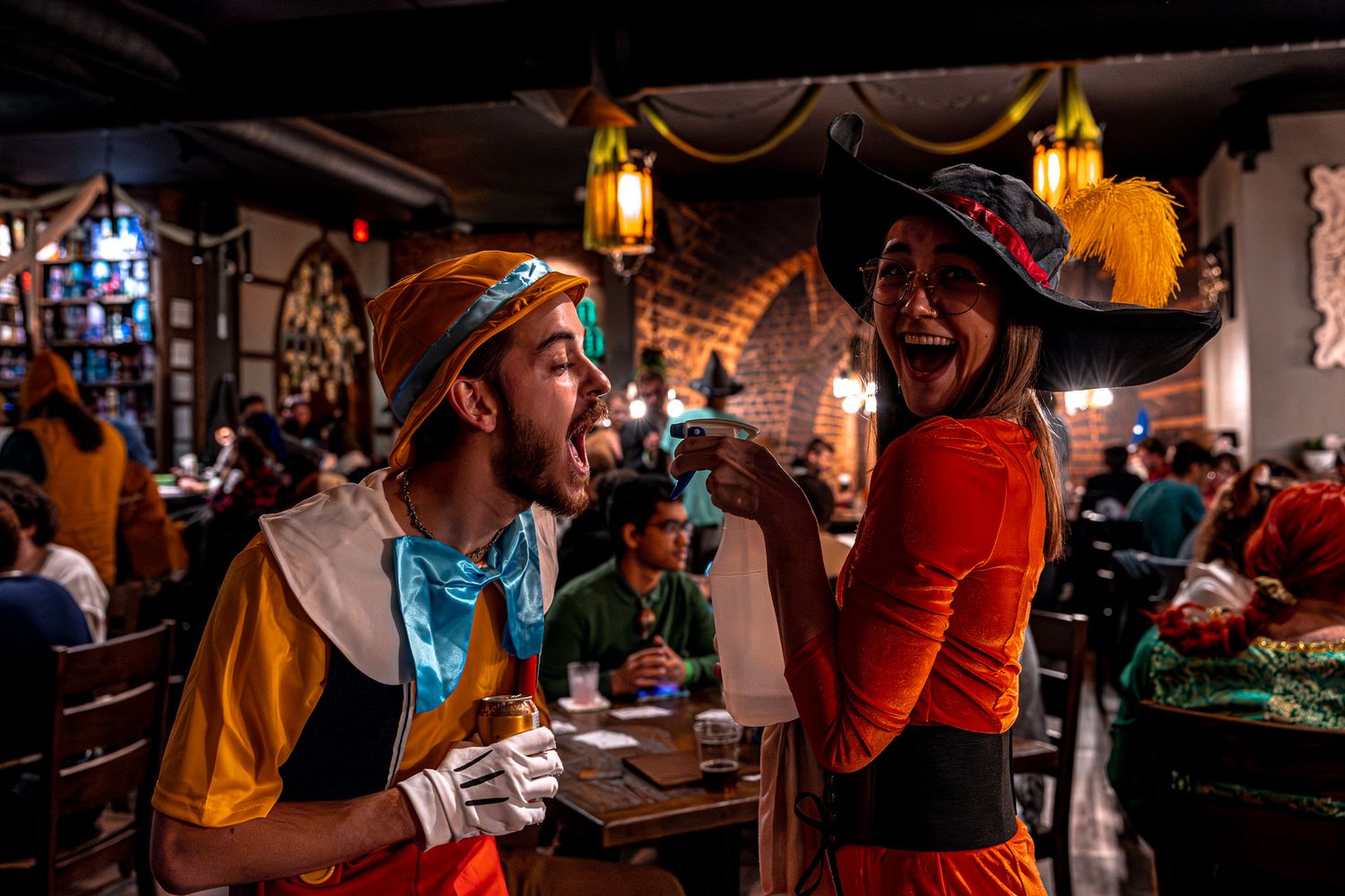 Two people dressed in colorful medieval festival costumes laugh and interact at a dimly lit indoor event venue.
