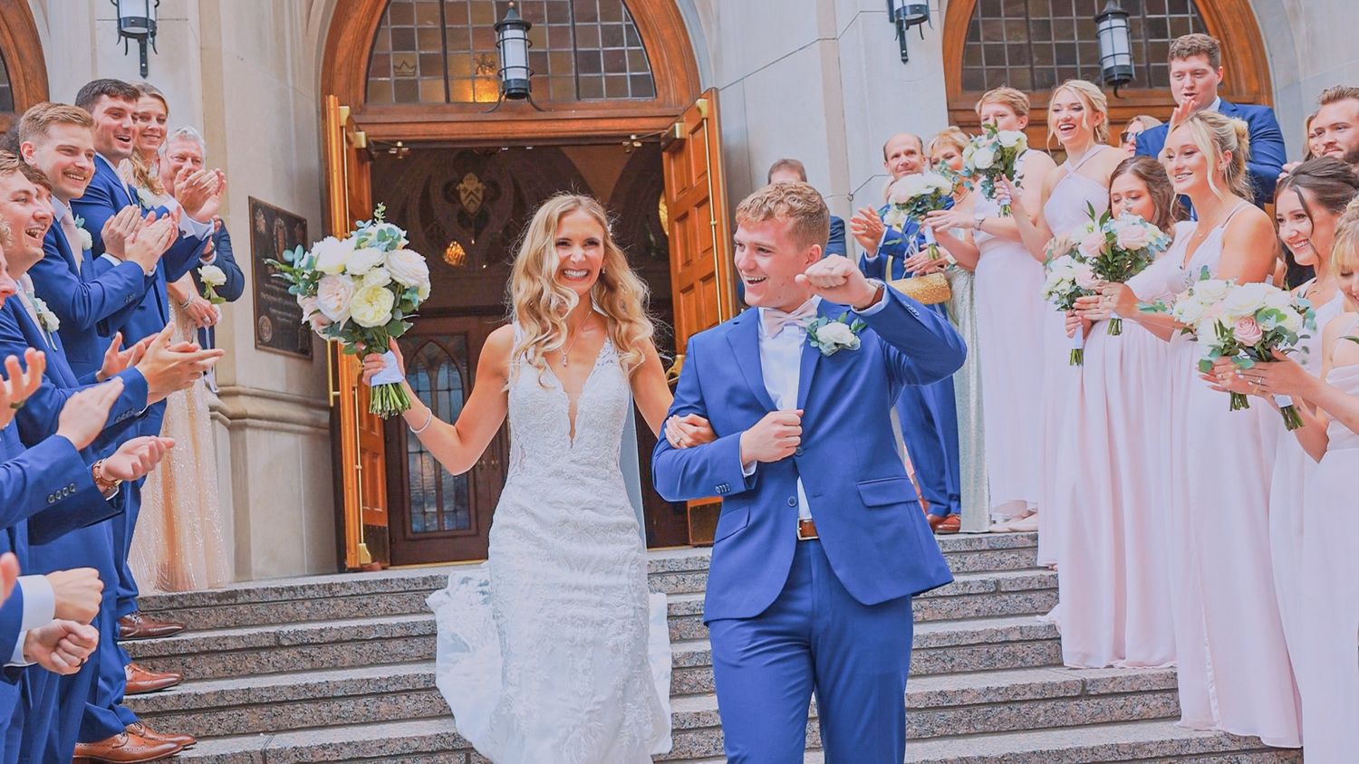 Wedding party celebrates on church steps as newlyweds in blue suit and white gown exit the ceremony.