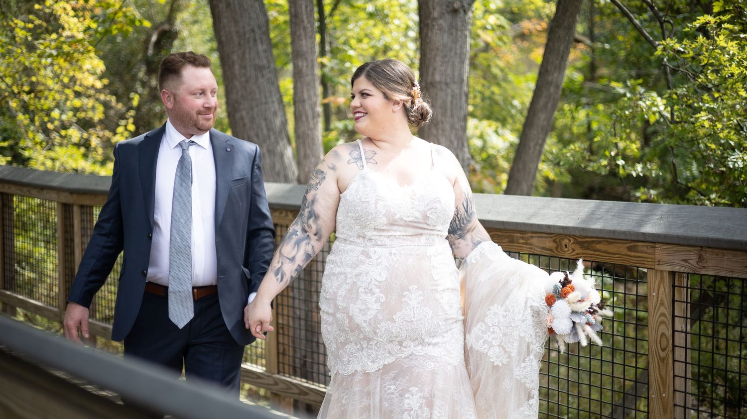 A couple walks hand in hand on a wooden bridge surrounded by autumn trees while wearing formal attire.