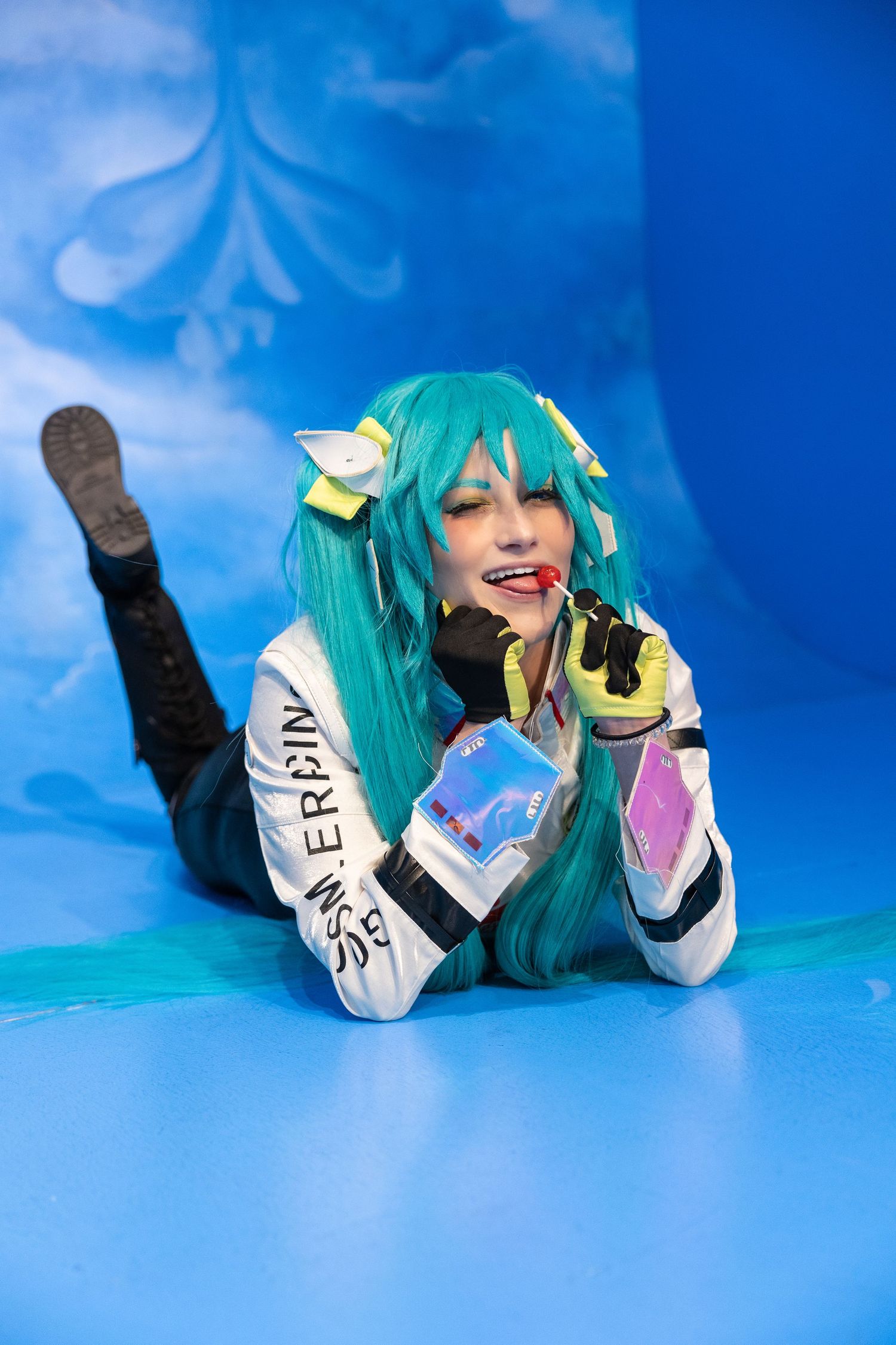 A cosplayer in a turquoise wig with yellow bows lies on a blue backdrop while holding a prop phone and smiling.