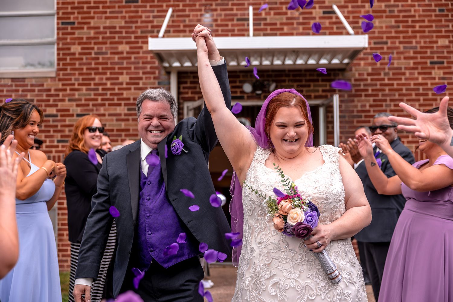 Newlyweds celebrate outside brick church entrance as guests throw confetti in purple and lavender wedding theme.