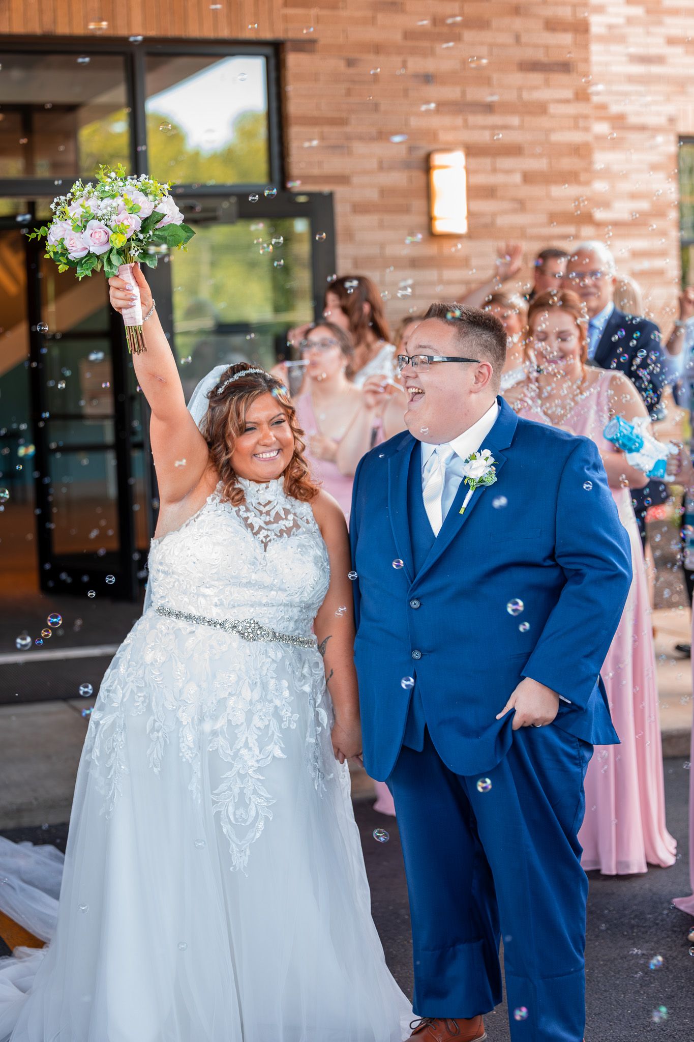 A joyful wedding celebration with bubbles floating in the air as the couple exits the venue in navy suit and white gown.