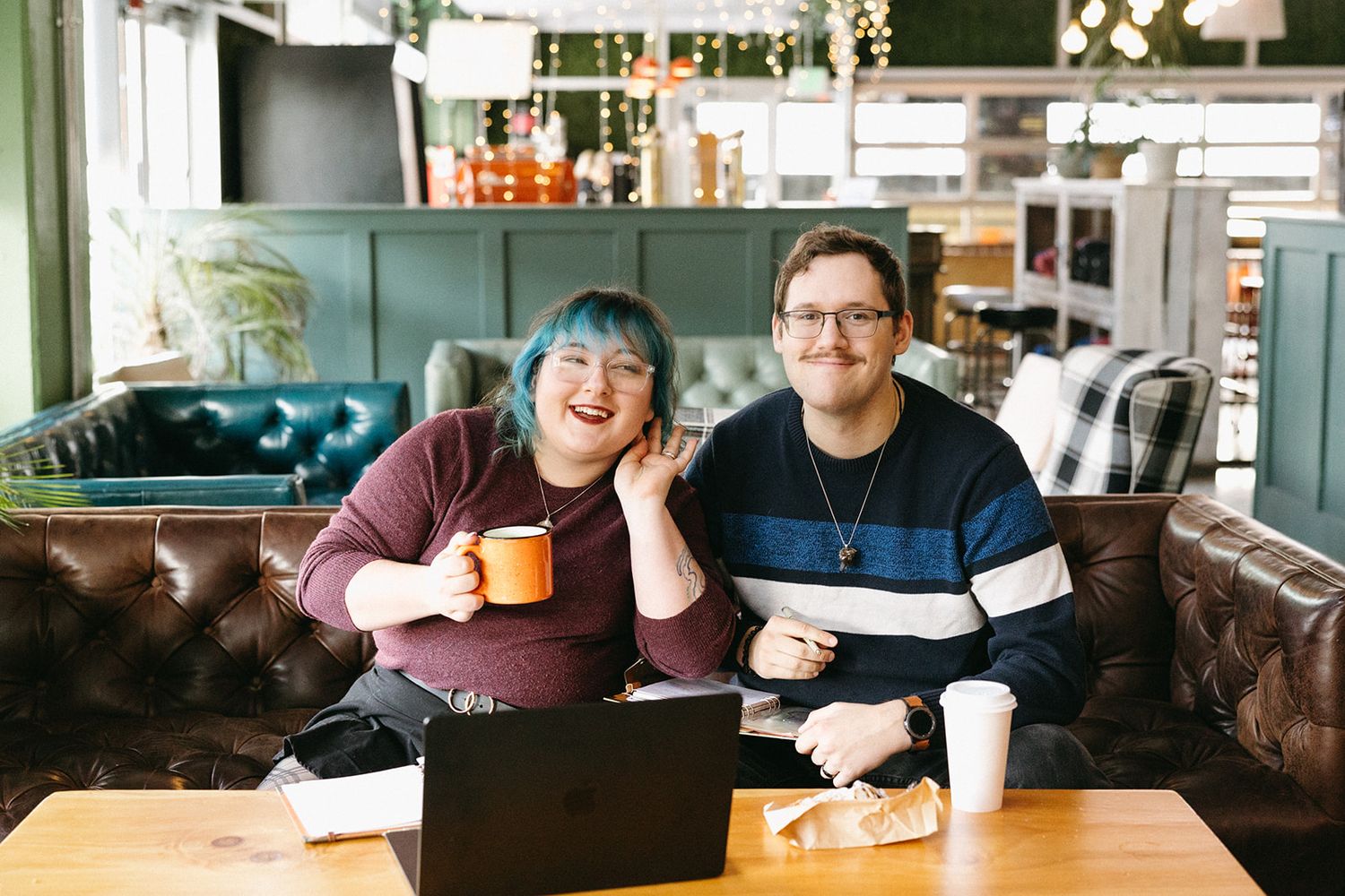 Two people sit at a coffee shop table with laptop and coffee mugs, smiling while working together in a cozy setting.
