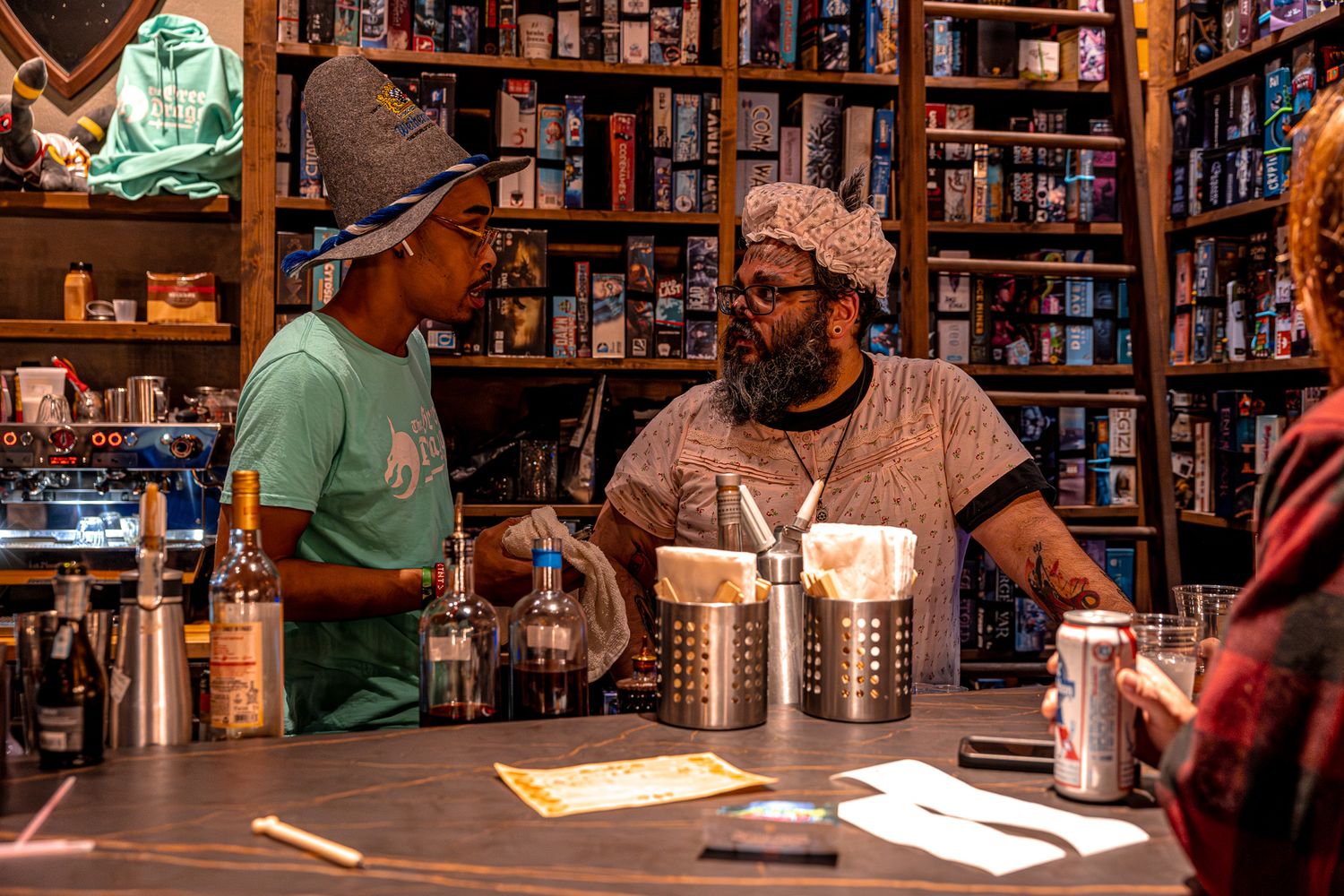 Two people engage in conversation at a bar counter with bookshelves visible in the background.