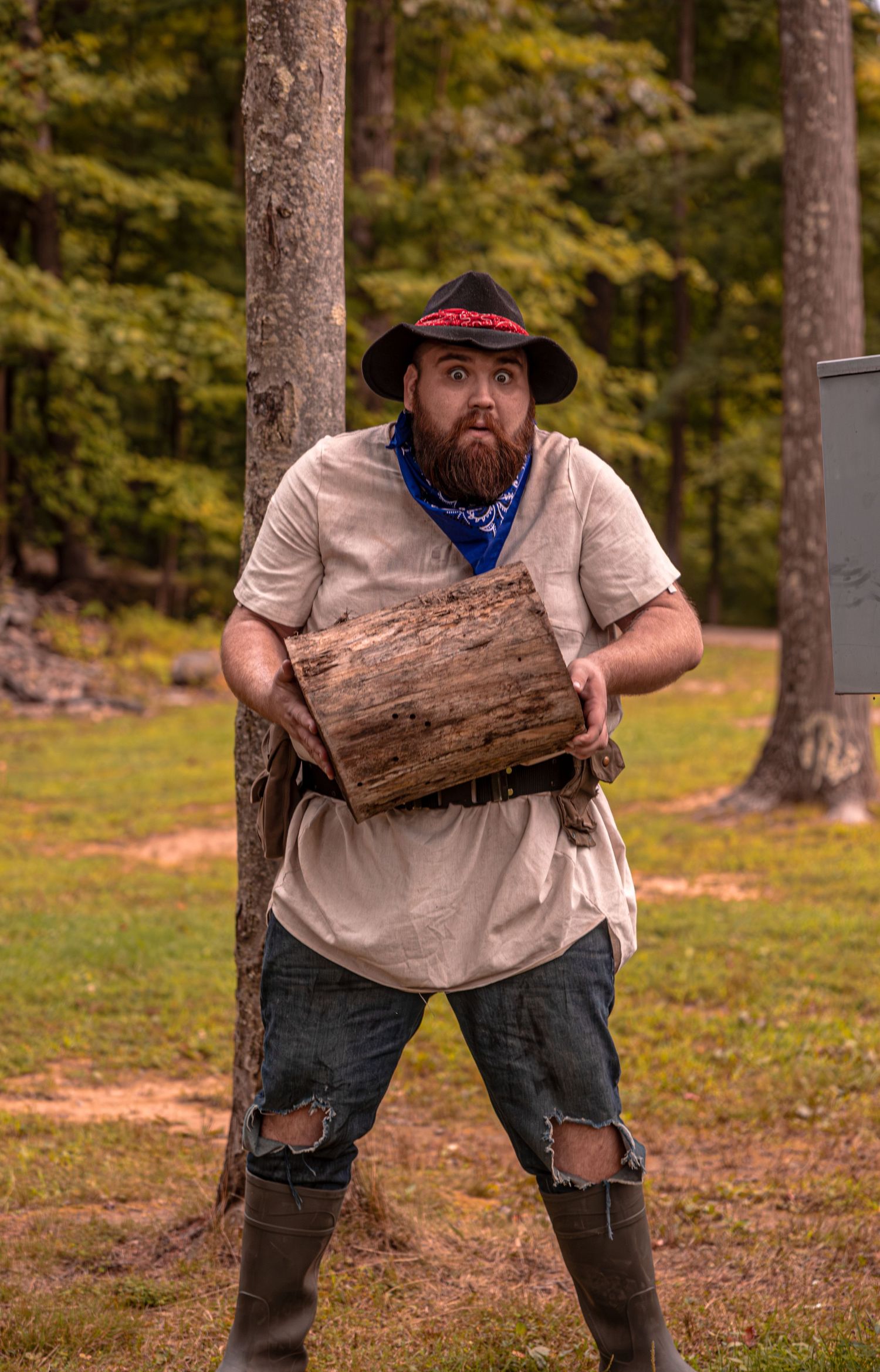 Someone in outdoor gear carries a large log in a wooded setting.