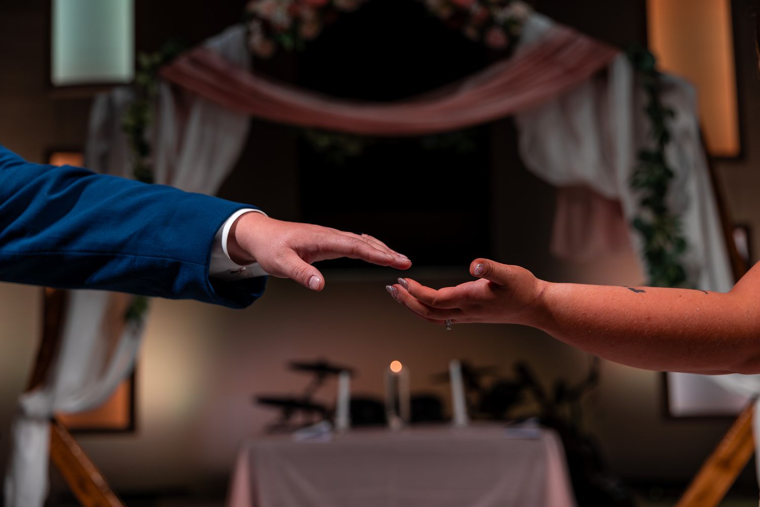 Two hands reaching towards each other during a wedding ceremony with decorative backdrop visible in background.