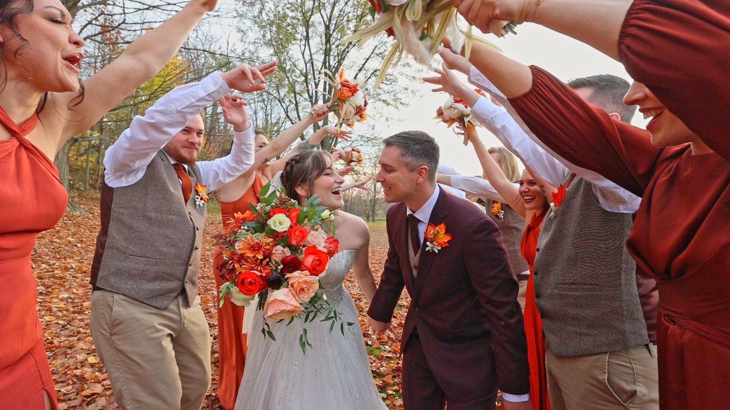 A couple shares a tender moment amidst orange autumn leaves while wedding party members throw petals in celebration.