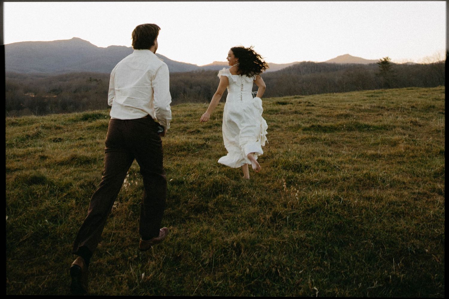 A couple runs joyfully through a grassy mountain meadow at sunset, with the bride's white dress flowing in the wind.