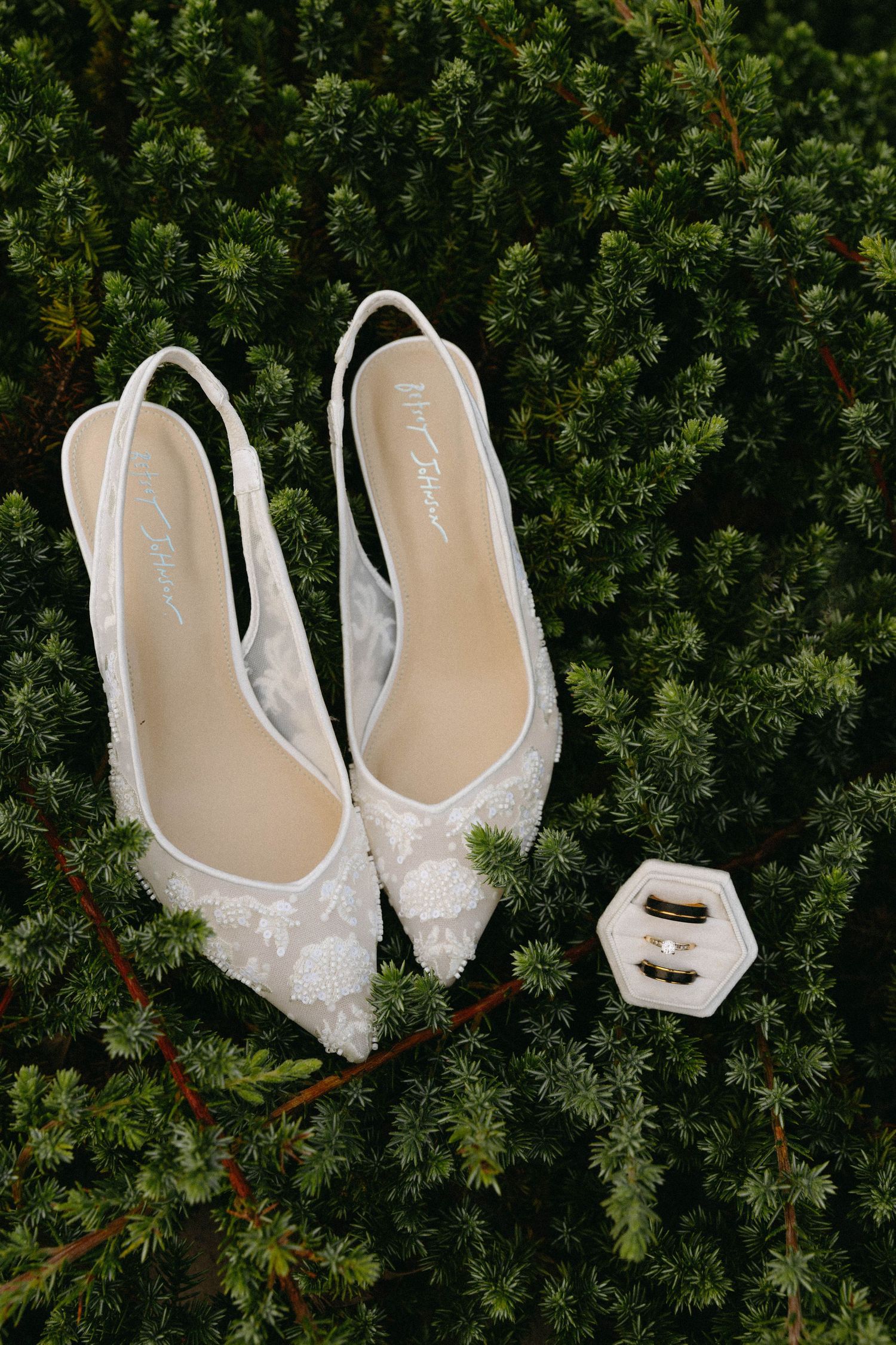 Elegant white lace bridal shoes displayed against lush green evergreen foliage background.