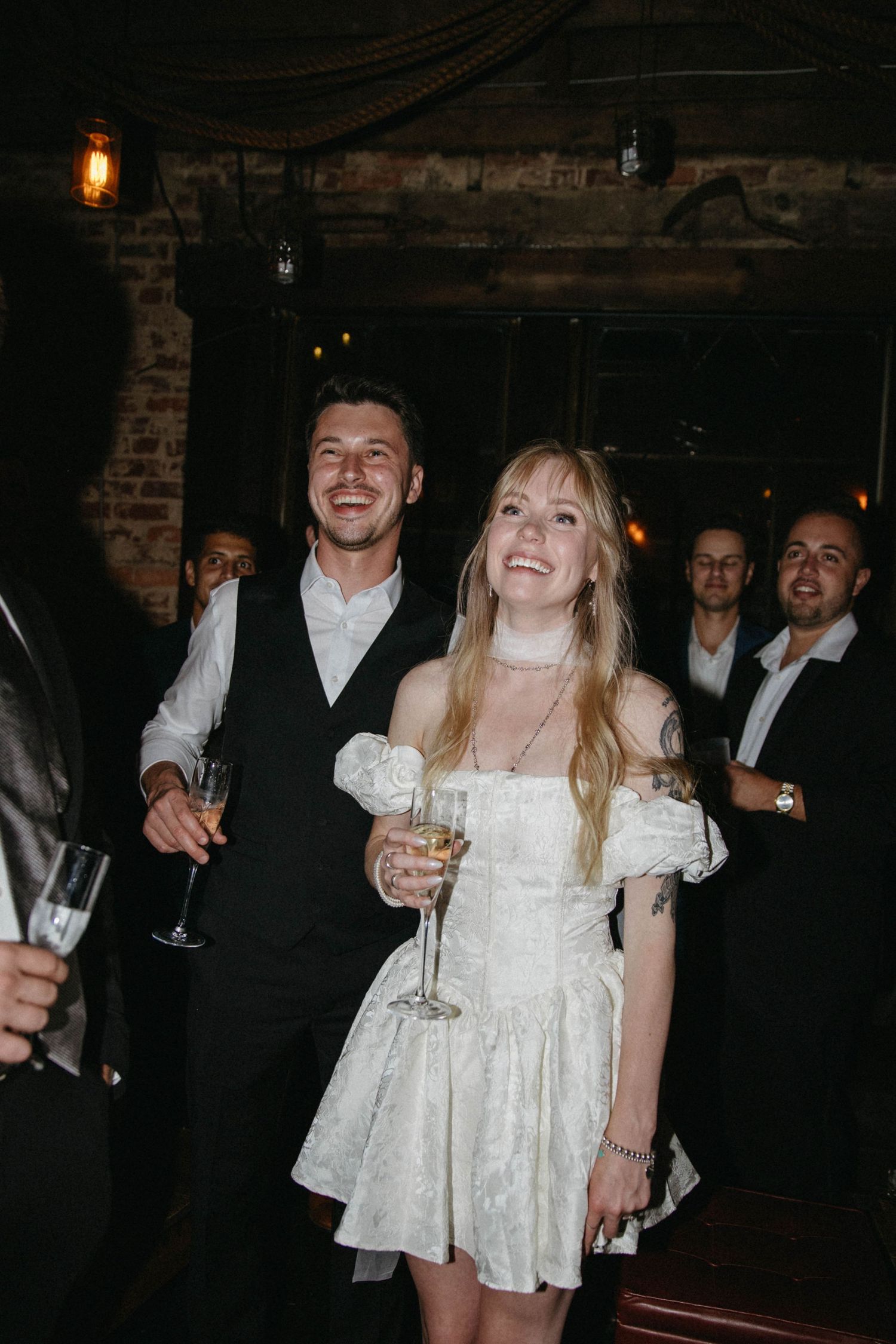 Couple celebrating at a dimly lit venue with guests laughing in the background.