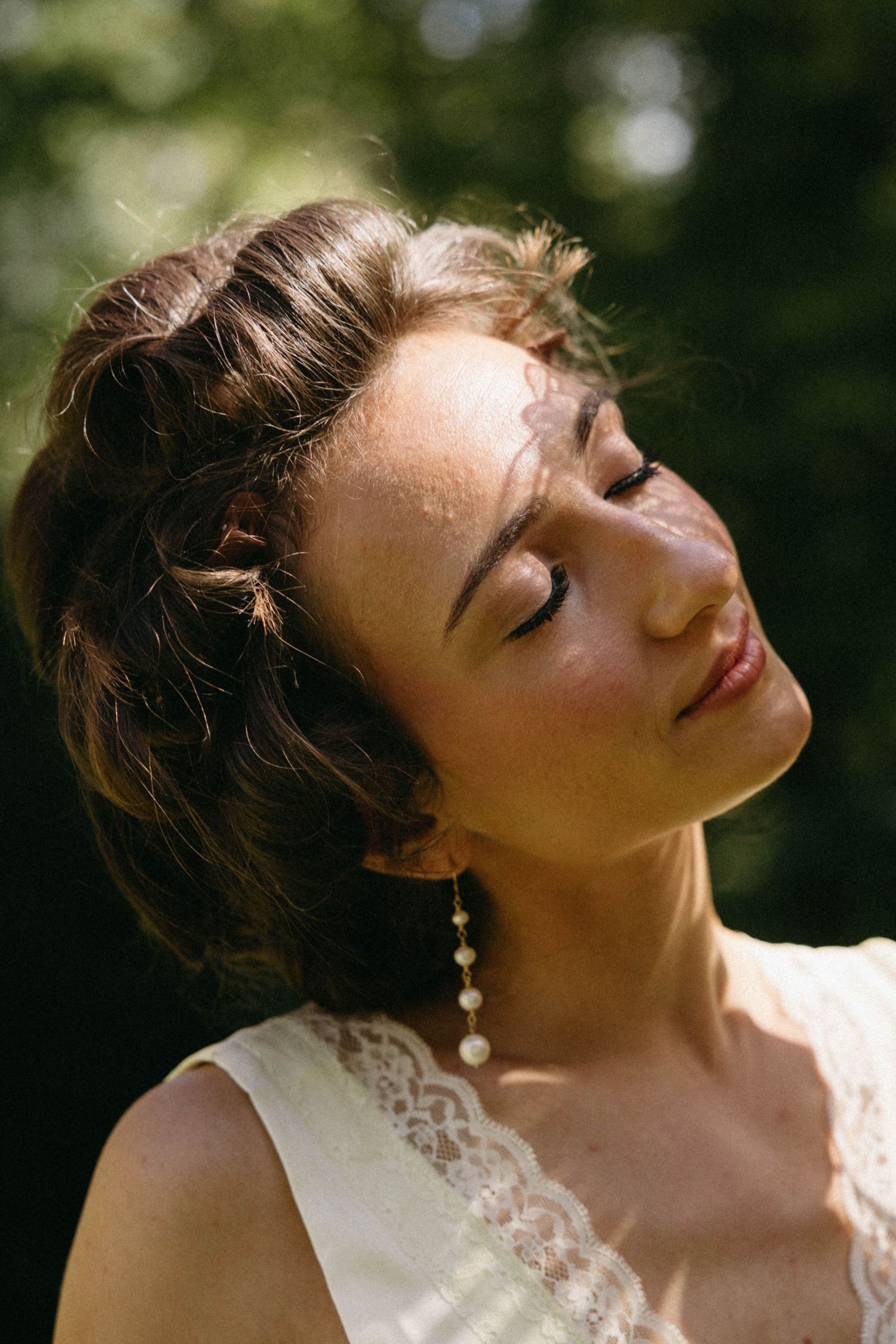 A dreamy portrait in sunlight shows a person wearing pearl earrings and a lace dress with eyes closed in peaceful repose.