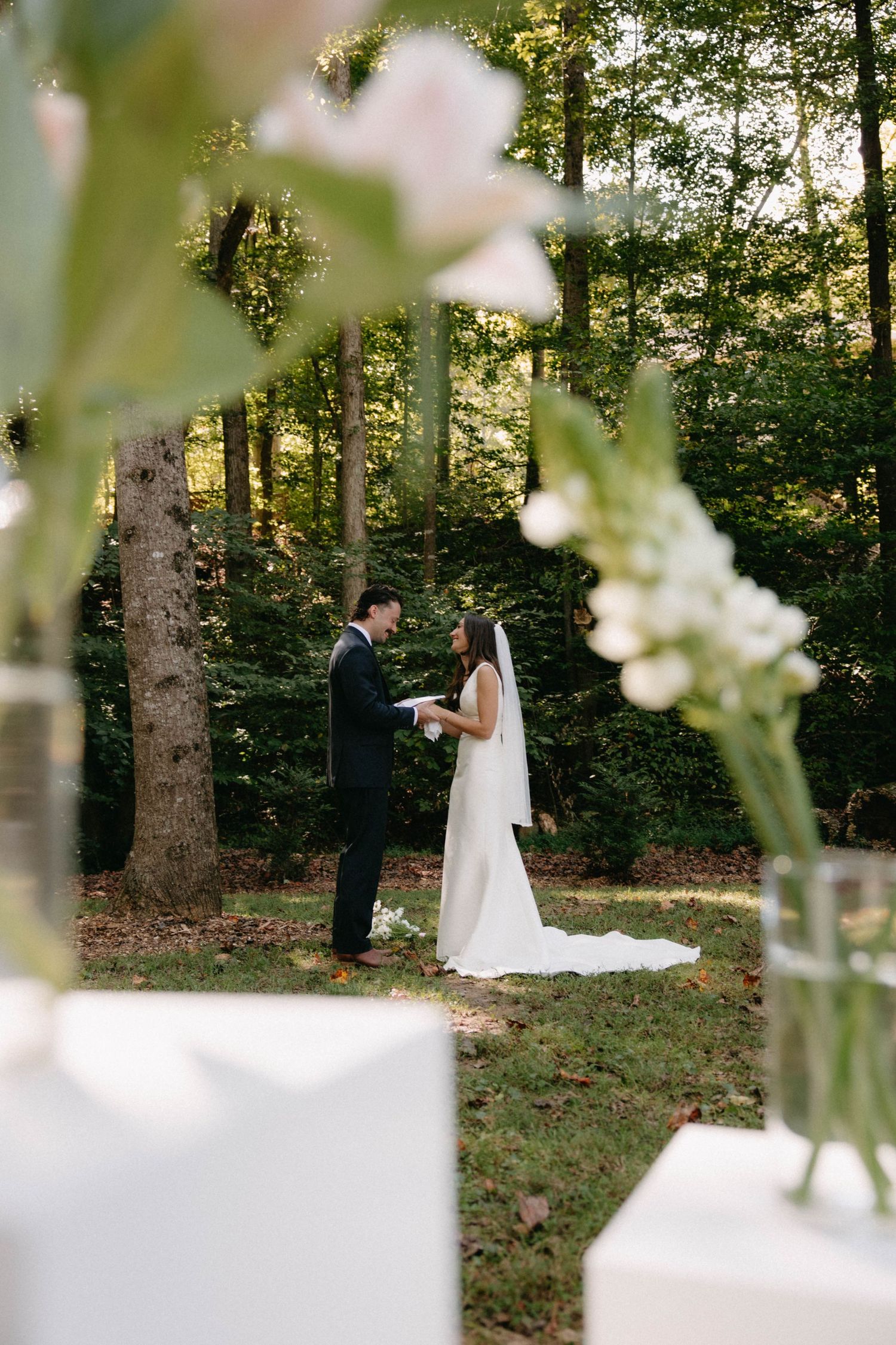 A romantic outdoor wedding ceremony takes place in a wooded forest setting with white floral decorations at sunset.
