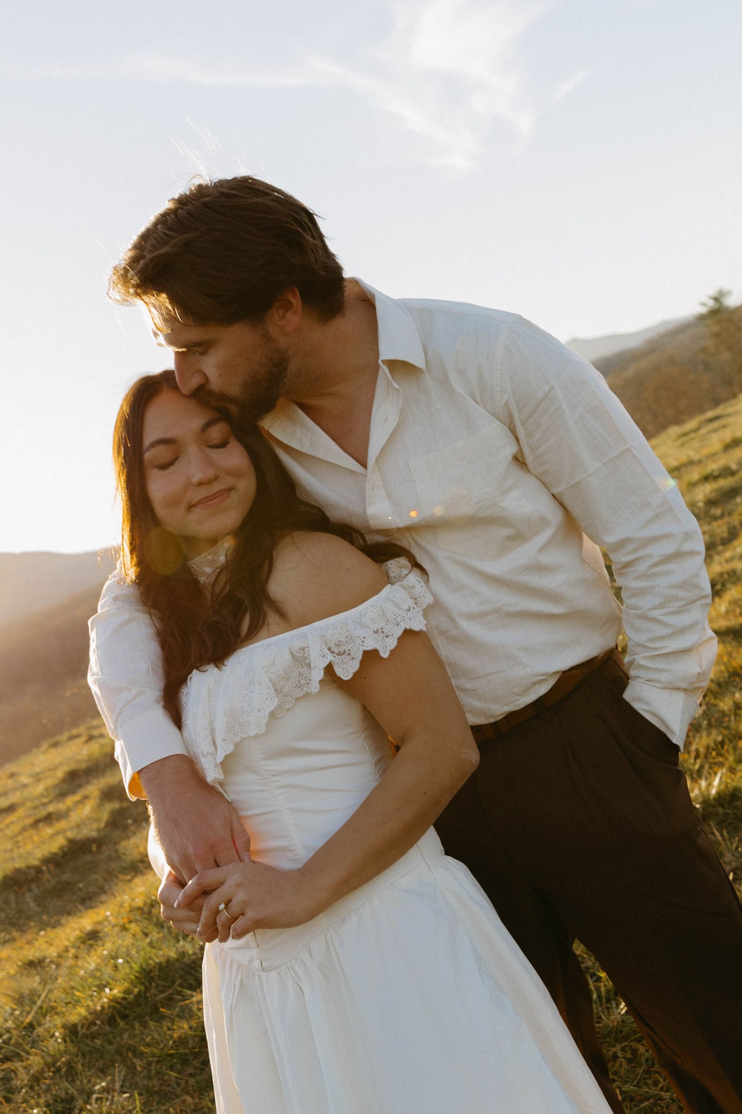 A romantic sunset wedding photo of a couple embracing on a hillside with golden light glowing behind them.