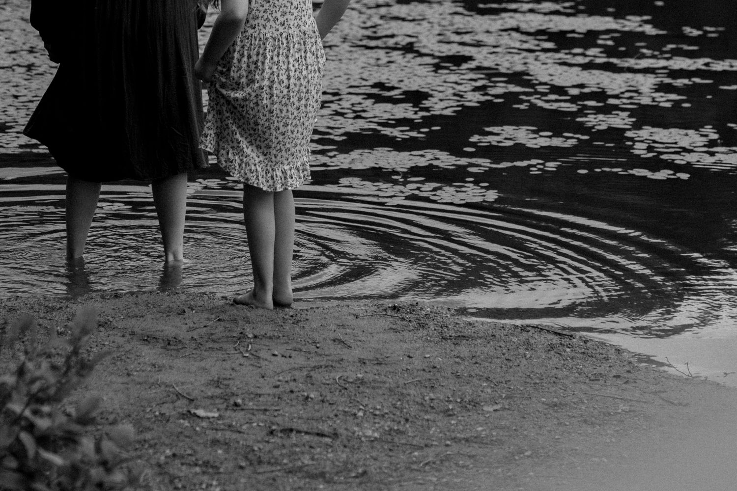 Black and white image of legs standing at water's edge creating ripples in the surface.
