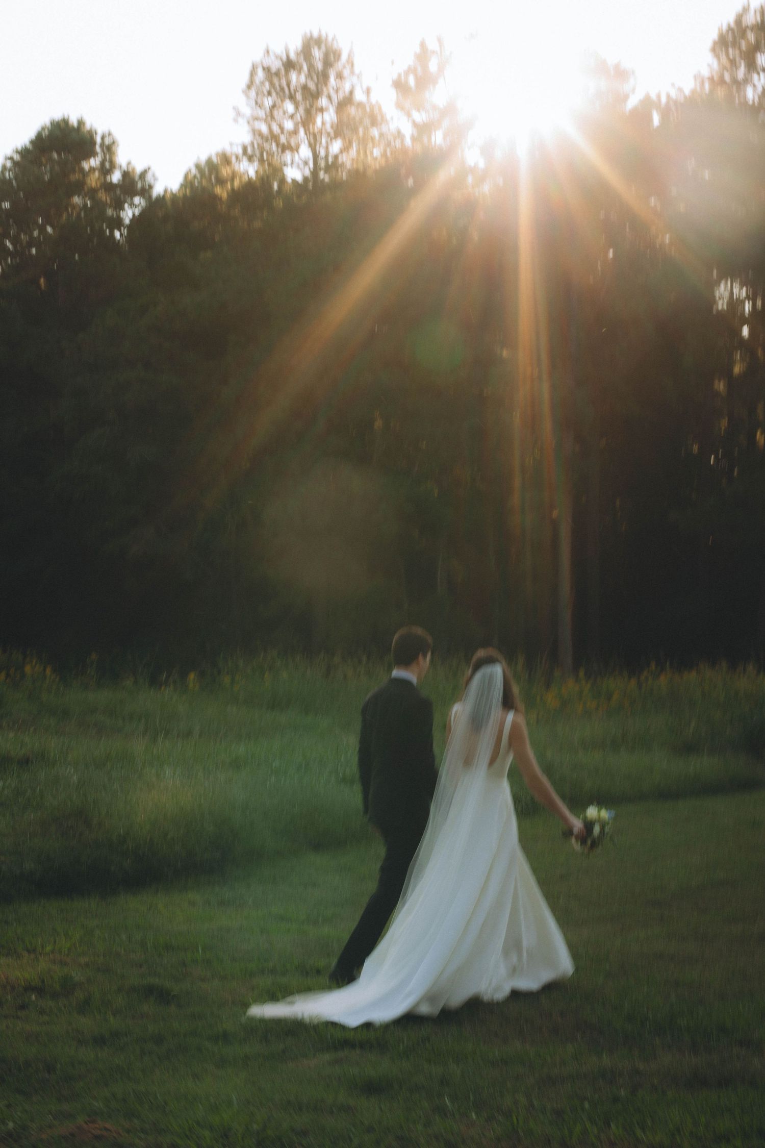 Couple walking through sunlit meadow at golden hour in wedding attire.