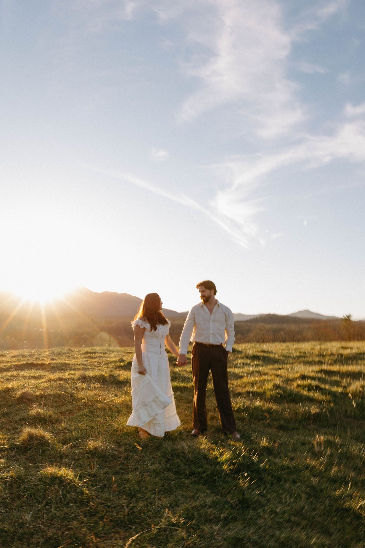 A couple walks together in a grassy meadow at sunset with mountains in the background.