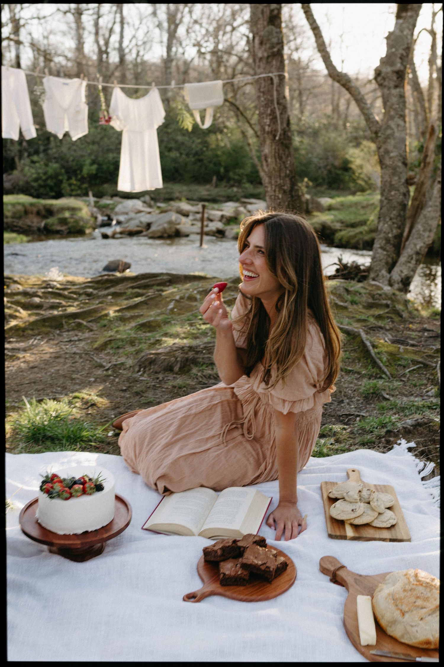 A picnic scene by a river with white blanket, bread, and books spread out on a sunny afternoon in nature.
