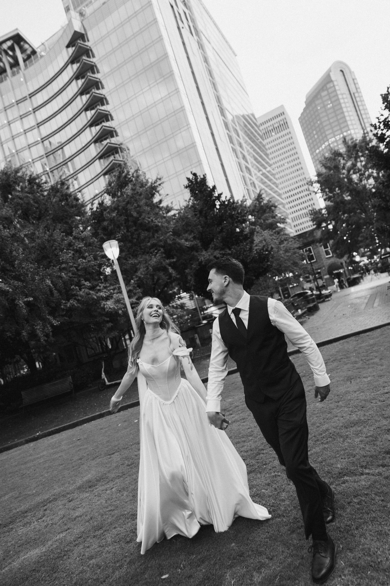 A couple in wedding attire take a playful walk together in front of modern city buildings in black and white photography.