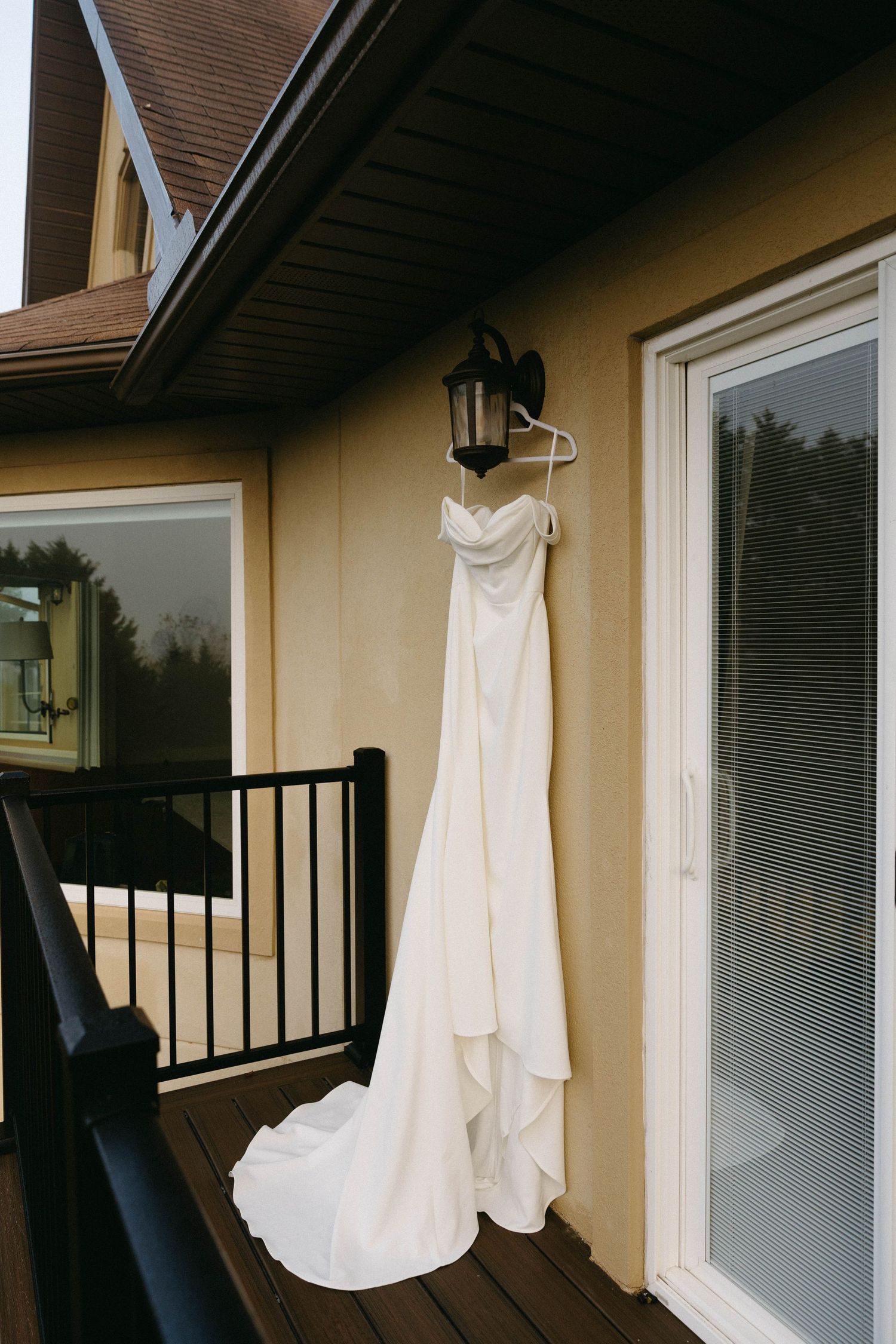 White wedding dress hanging on outdoor balcony railing against beige wall and windows at dusk.