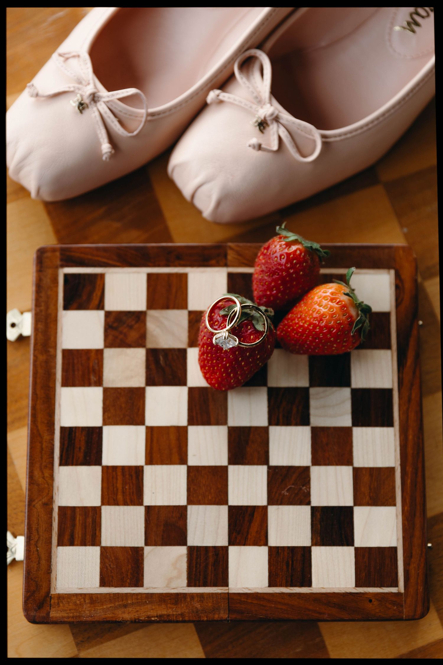 White ballet shoes and red strawberries arranged on wooden chess board.