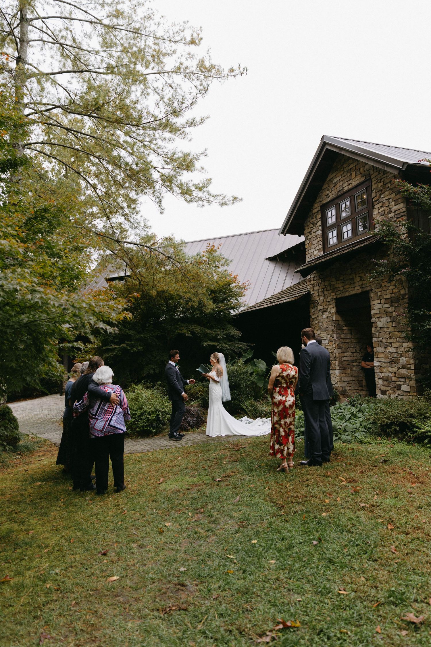 A small stone cabin on a hillside surrounded by autumn trees while a small group gathers outside.
