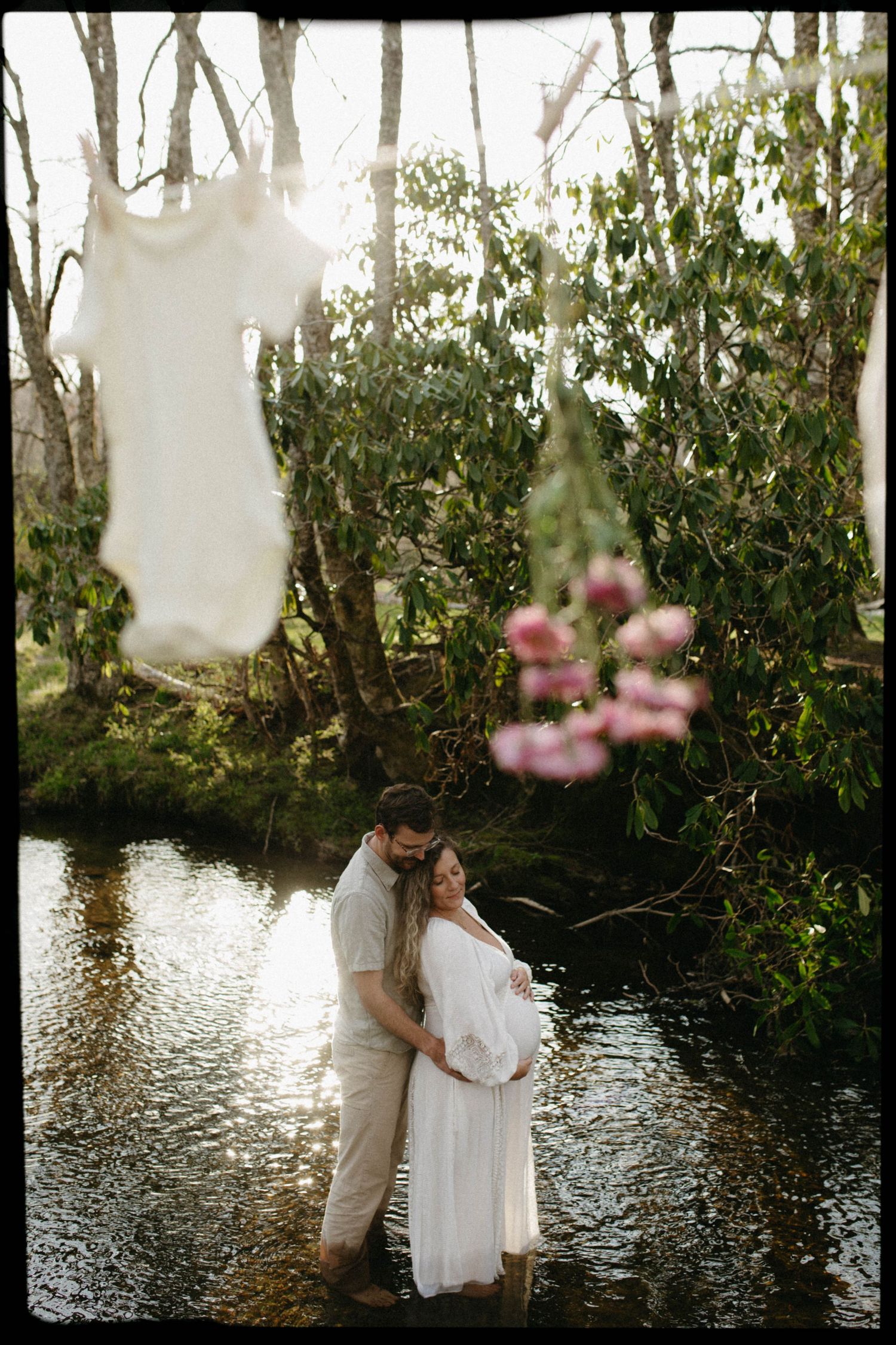 A romantic moment captured by a waterfront with flowering trees and soft sunlight filtering through the foliage.
