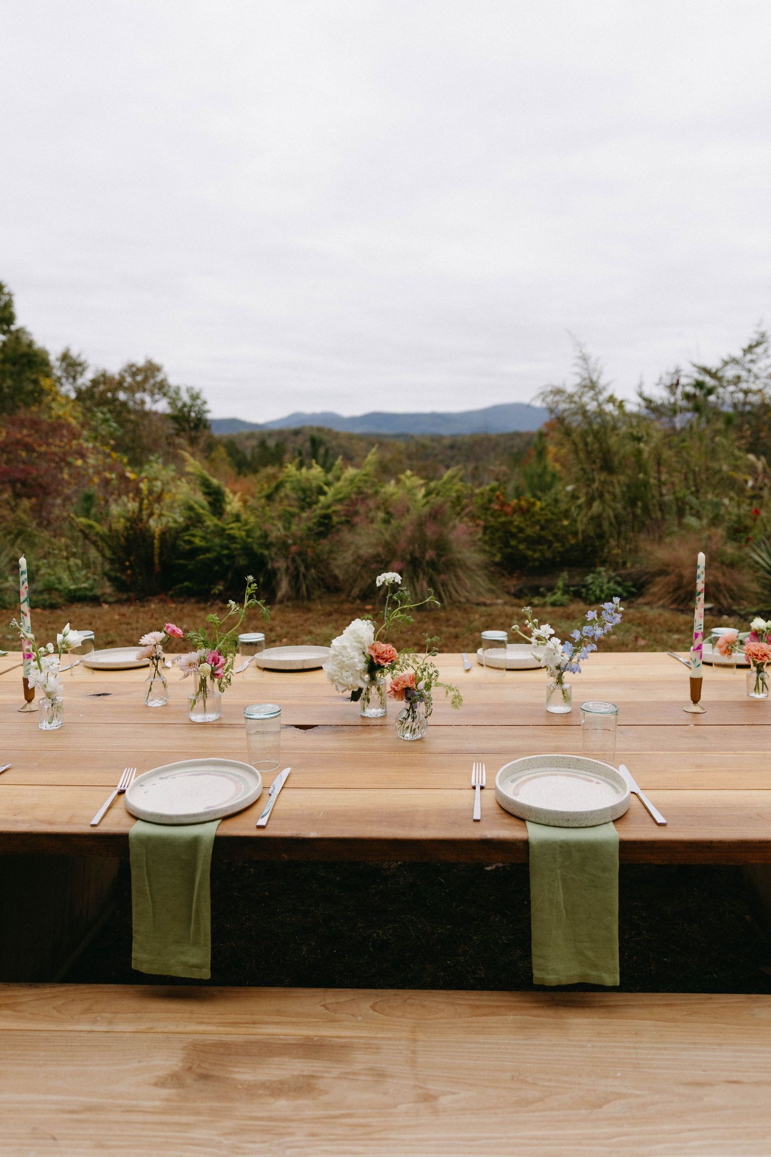 A rustic outdoor table setting with green napkins and small floral arrangements against a mountain backdrop.