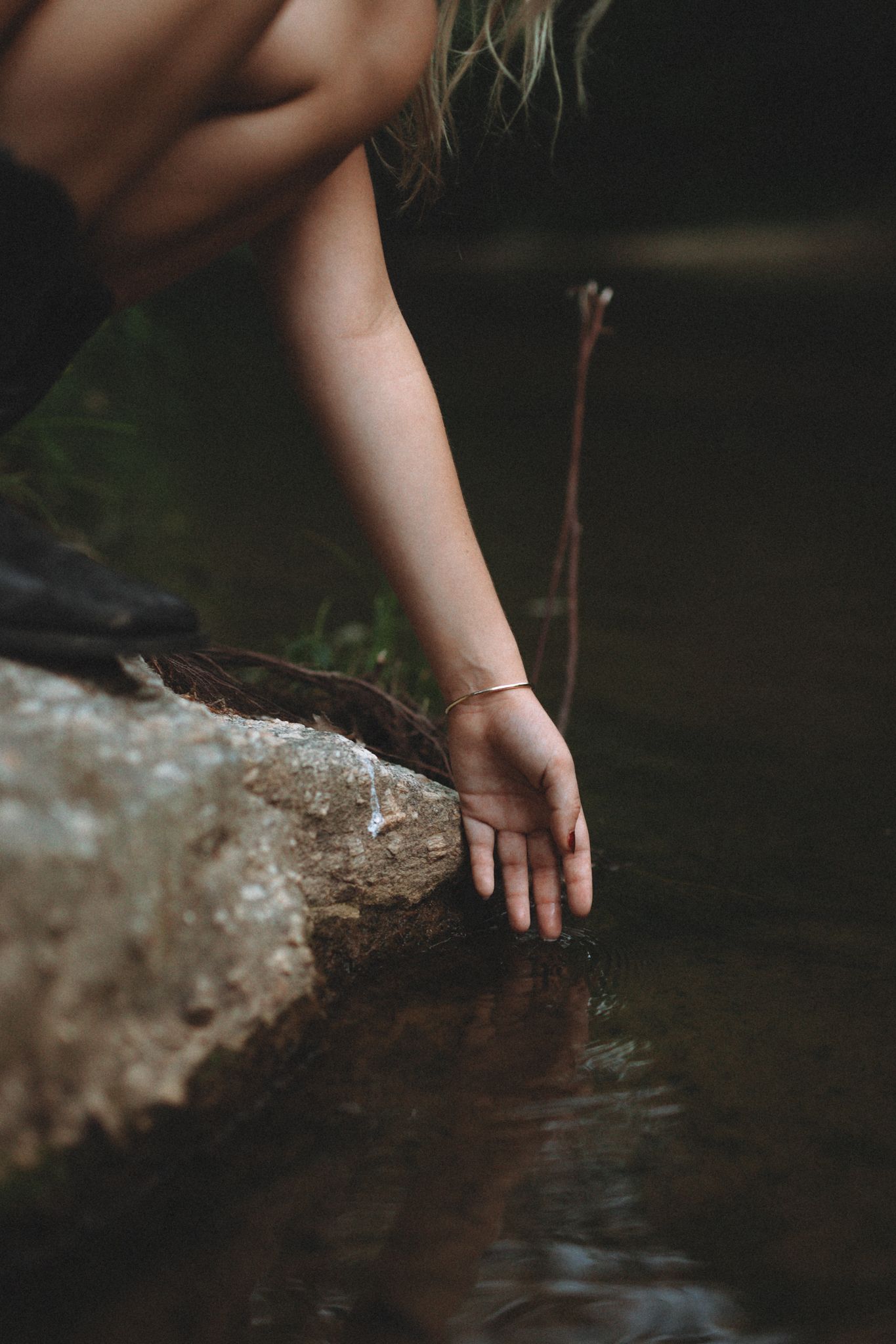 Artistic shot of hand with bracelet reaching down to touch water from rocky ledge.