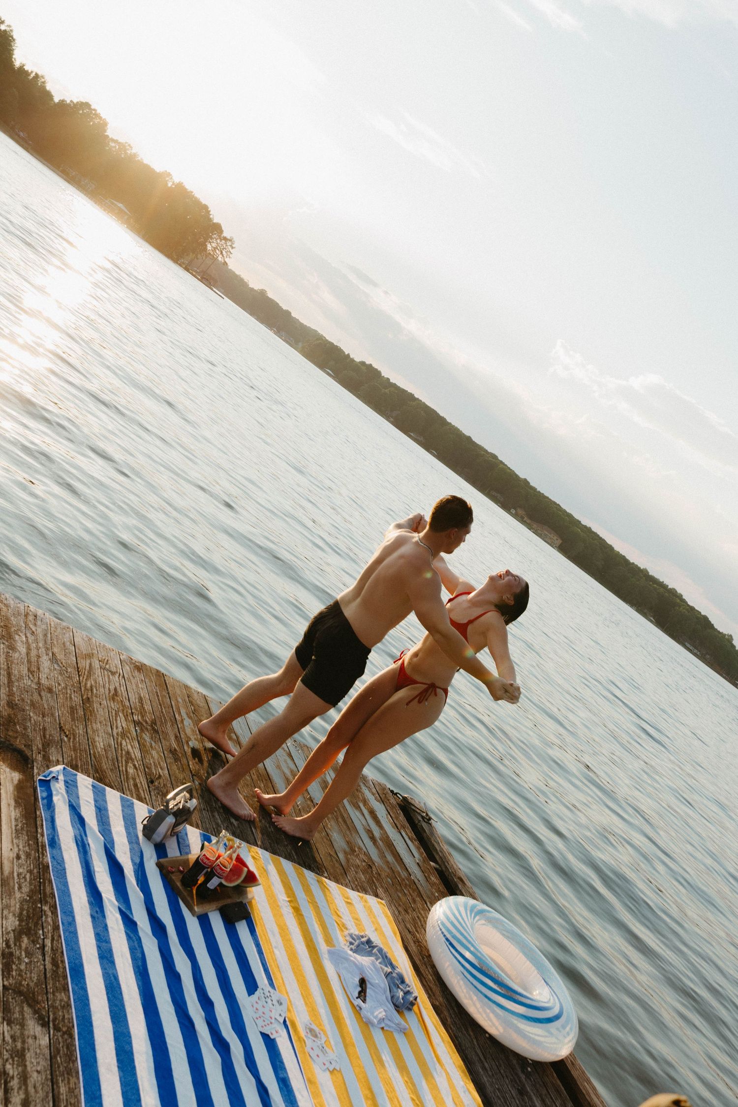 A couple jumps into a lake from a dock with beach towels and supplies laid out on a sunny summer day.
