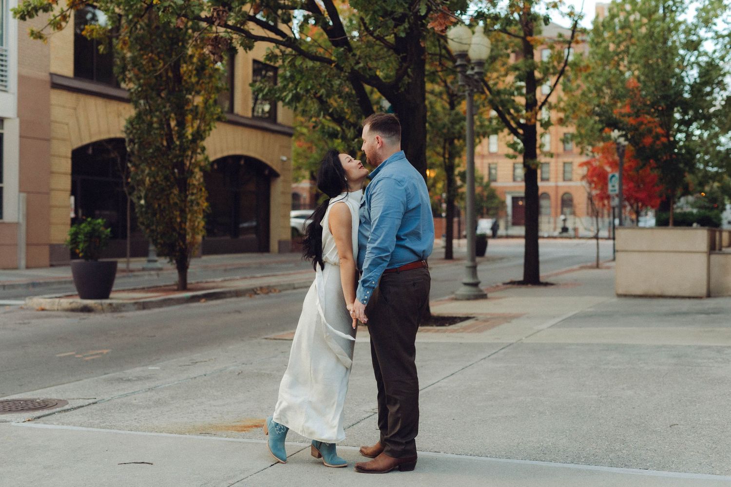 Romantic couple walks together outdoors along a city street with fall foliage in background.
