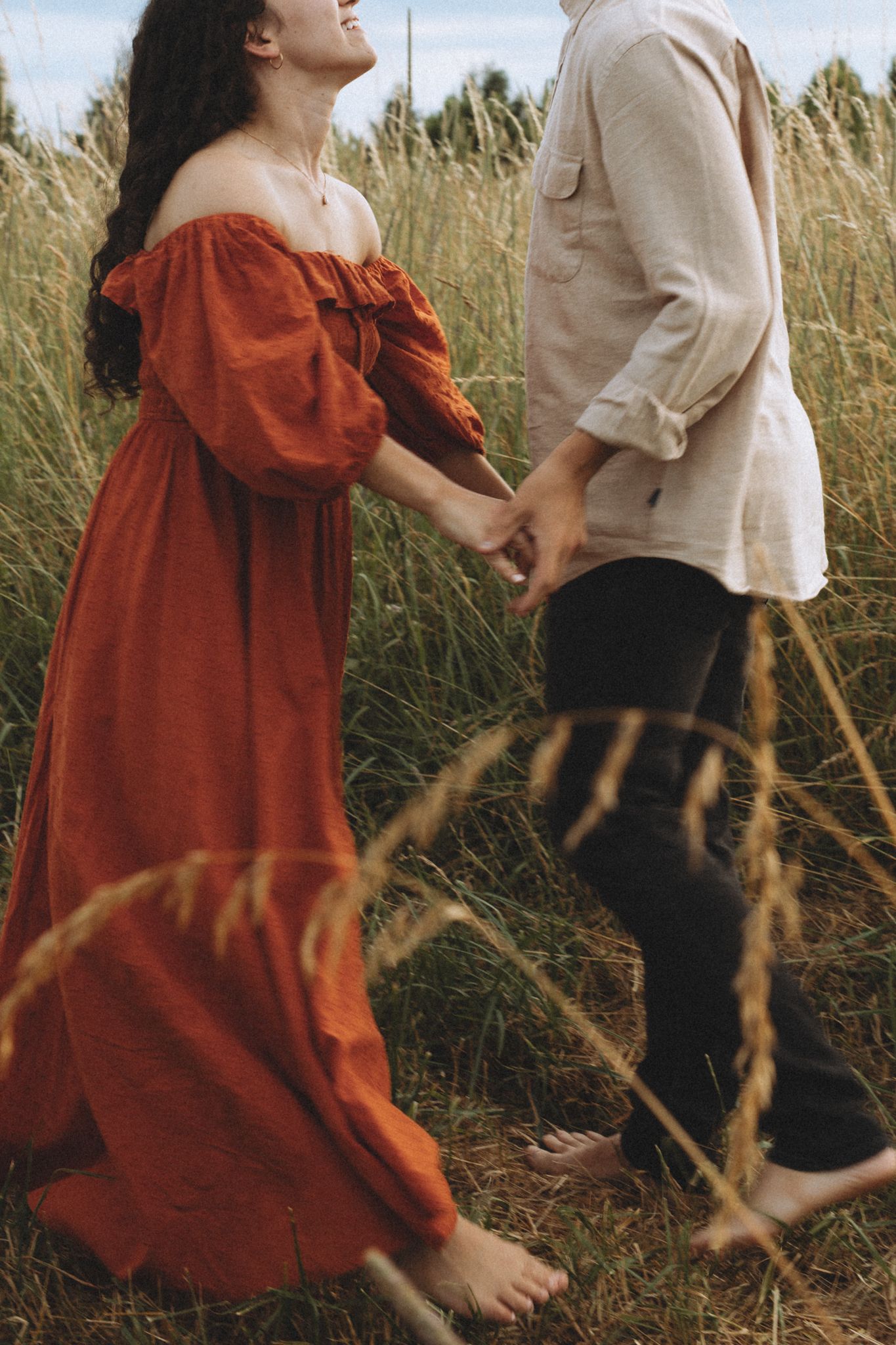 Two people walking through tall grass, one wearing a flowing red dress and the other in light clothing.