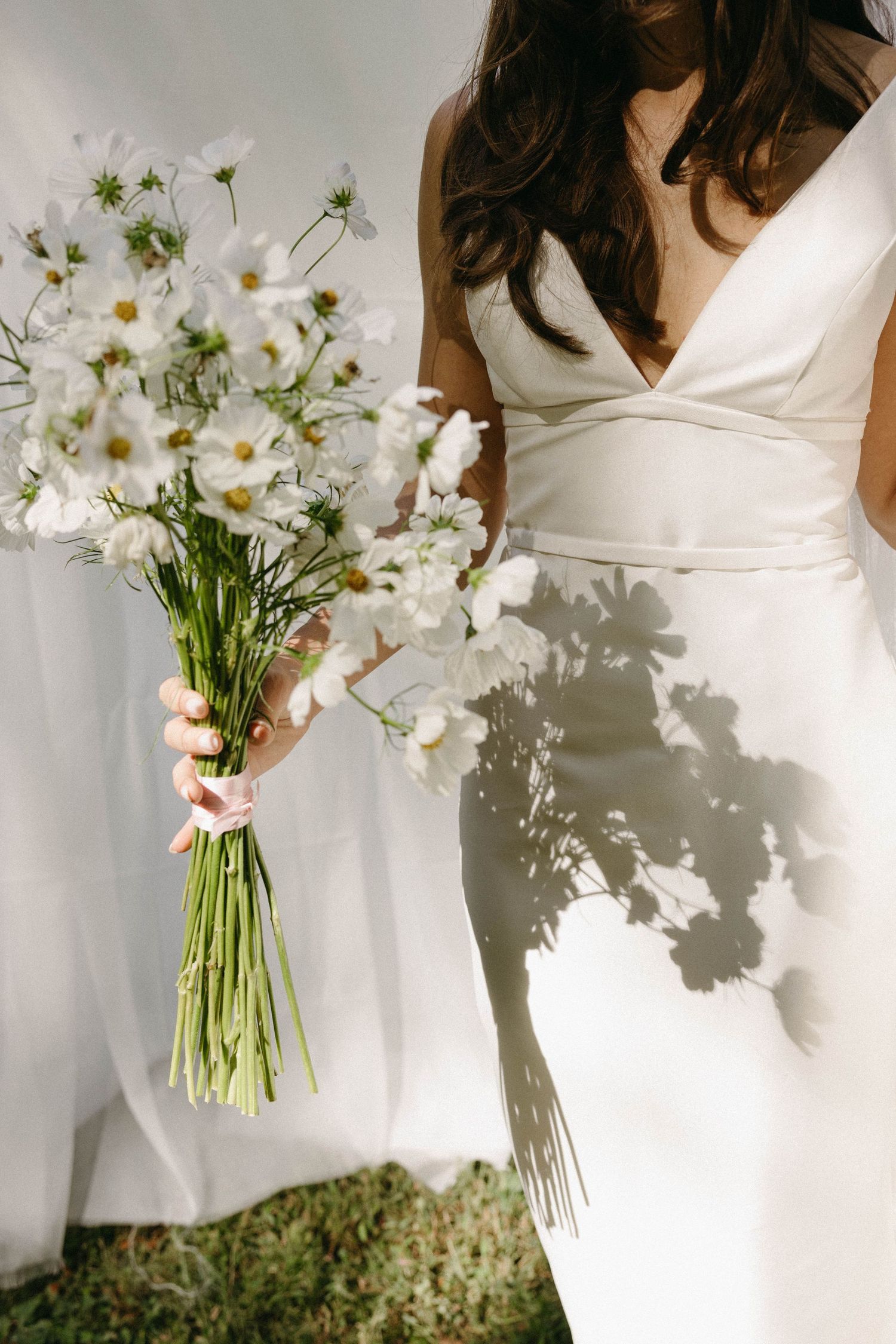 A white wedding dress and a delicate bouquet of white cosmos flowers casting shadows in natural light.