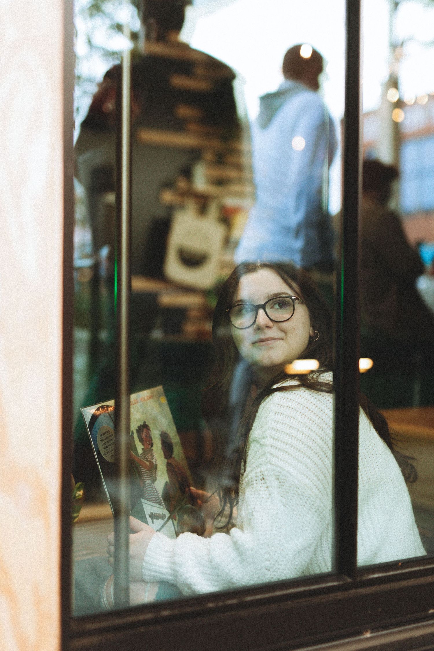 Someone sitting by a window in a white sweater, reflected in the glass pane.