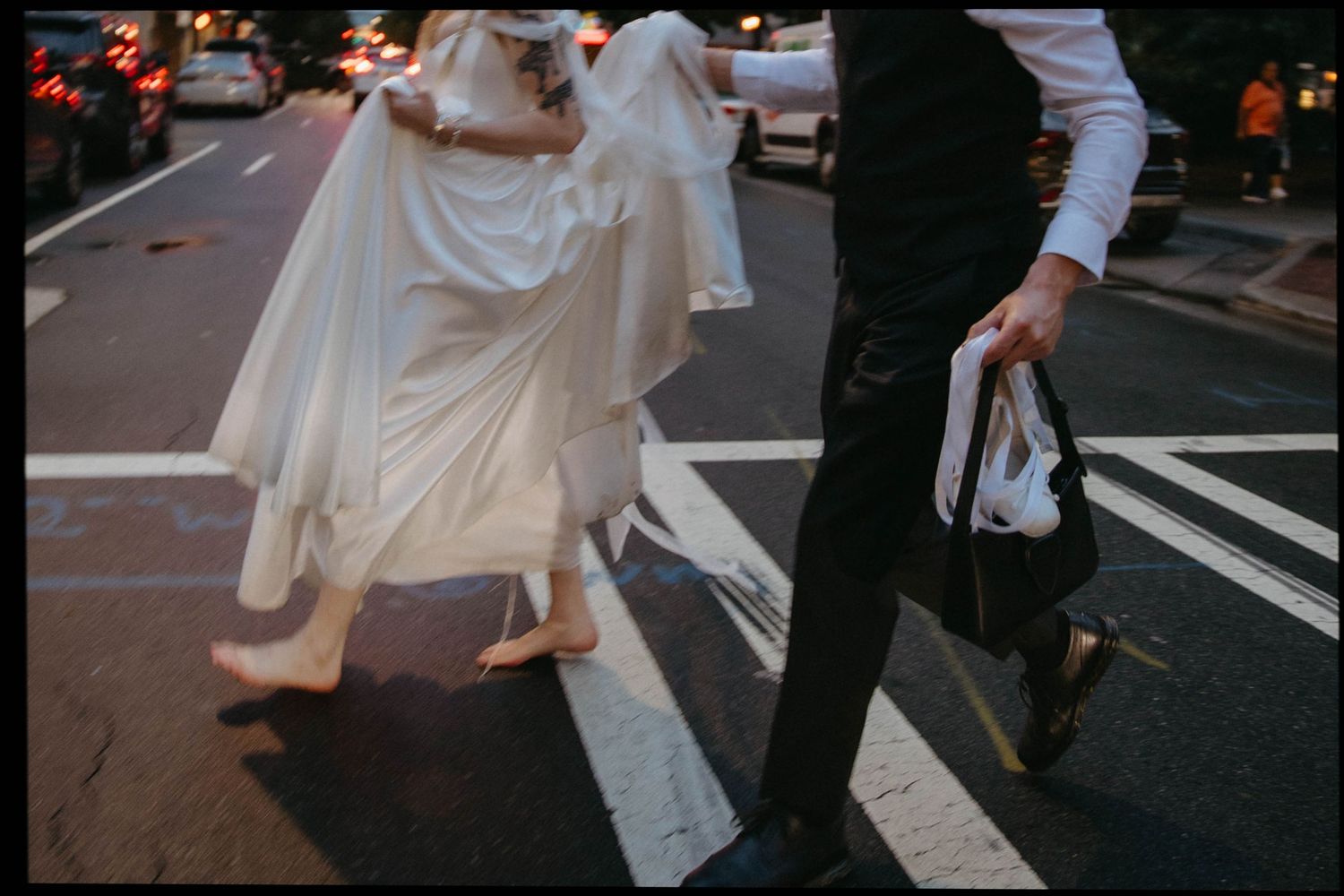 A bride in a flowing white dress and groom in dark suit cross a city street at dusk.