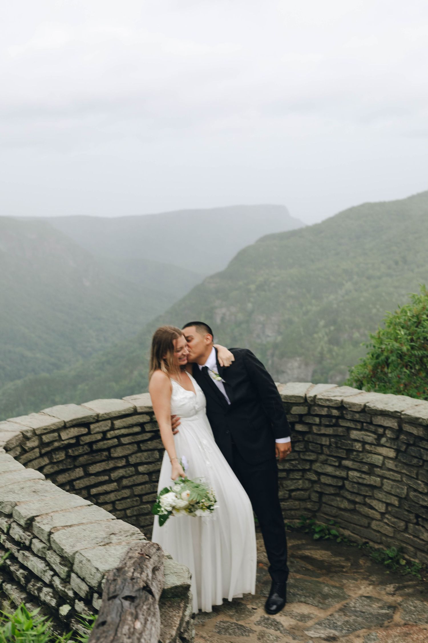 Wedding couple shares a romantic moment at a scenic mountain overlook with stone wall barrier on a misty day.