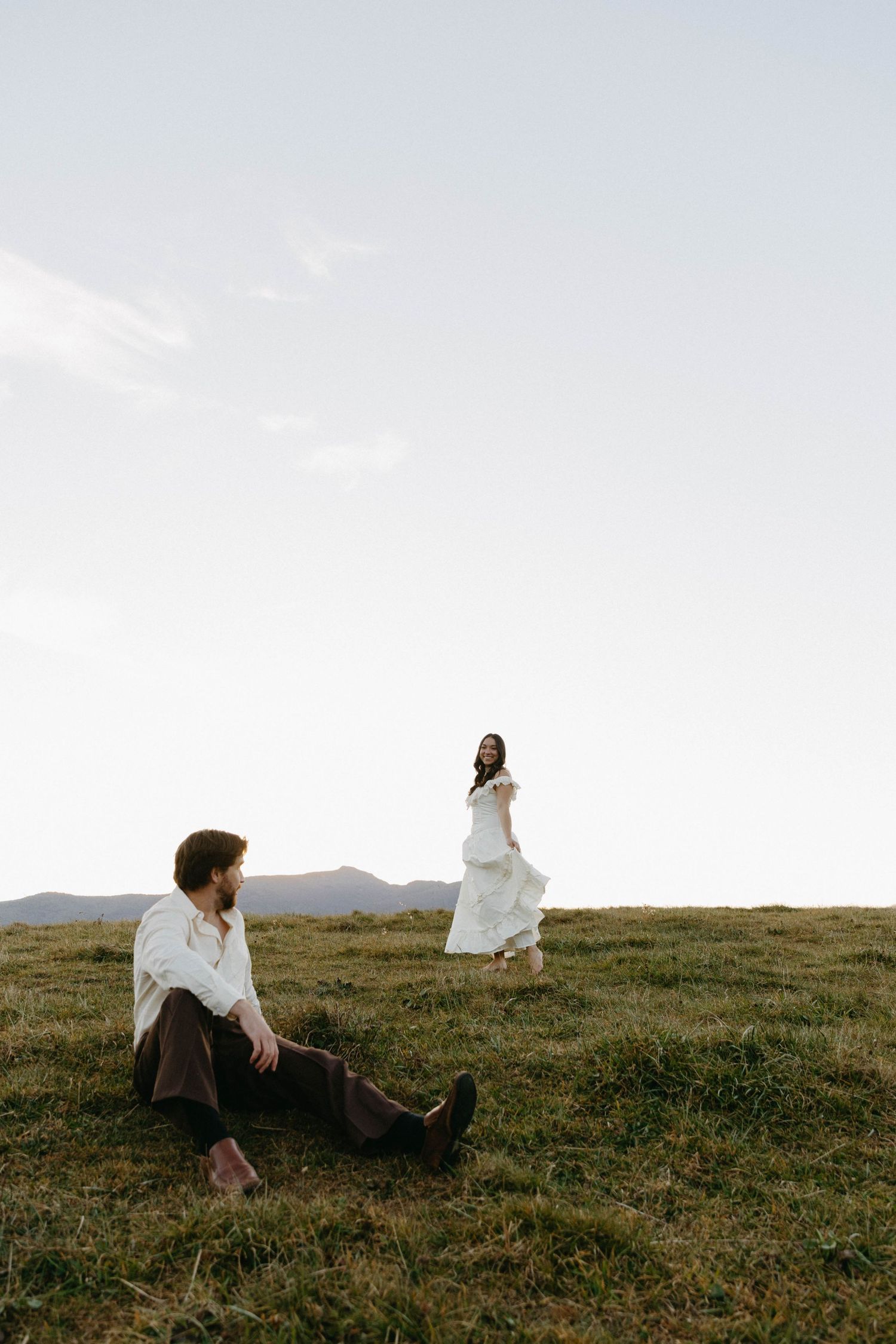 A couple on a grassy hilltop at sunset, with mountains in the background and soft lighting creating a romantic scene.
