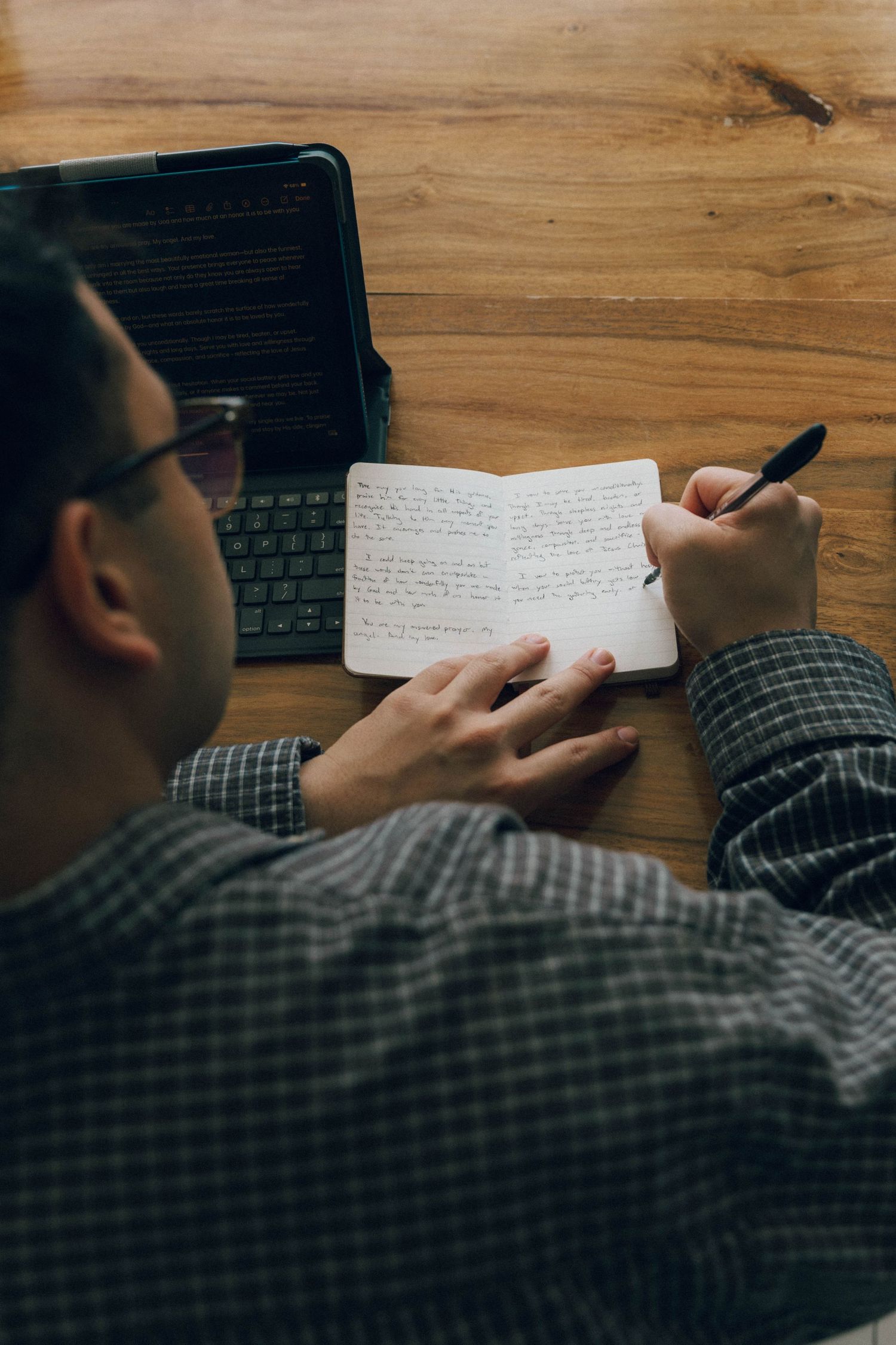 Person writing in notebook next to laptop on wooden desk during evening hours.
