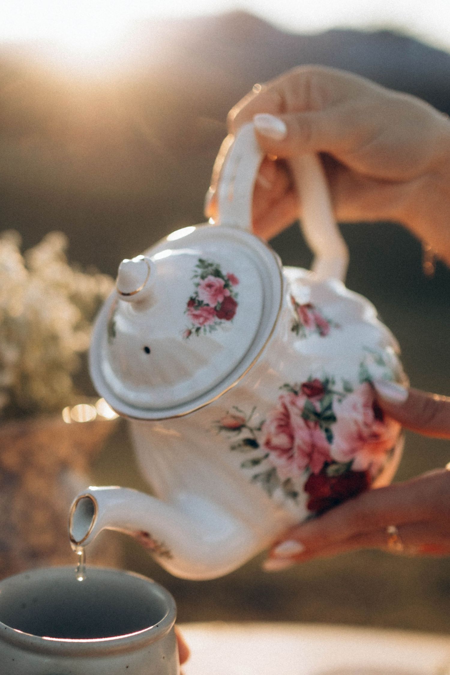 A floral teapot pouring hot tea with steam rising in golden sunlight.