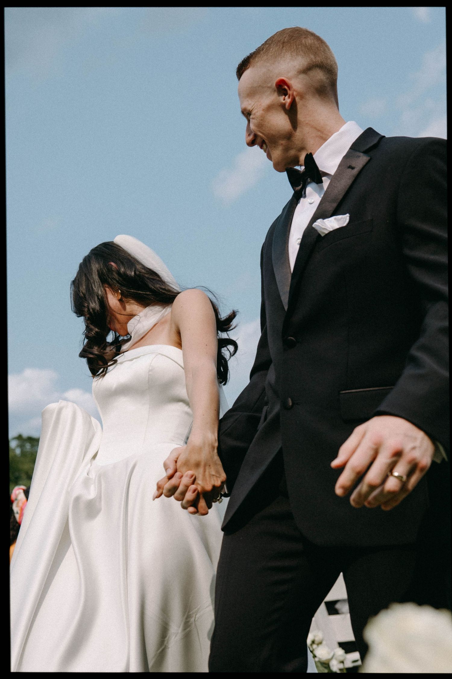 A couple in wedding attire hold hands and share a candid moment outdoors against a blue sky.
