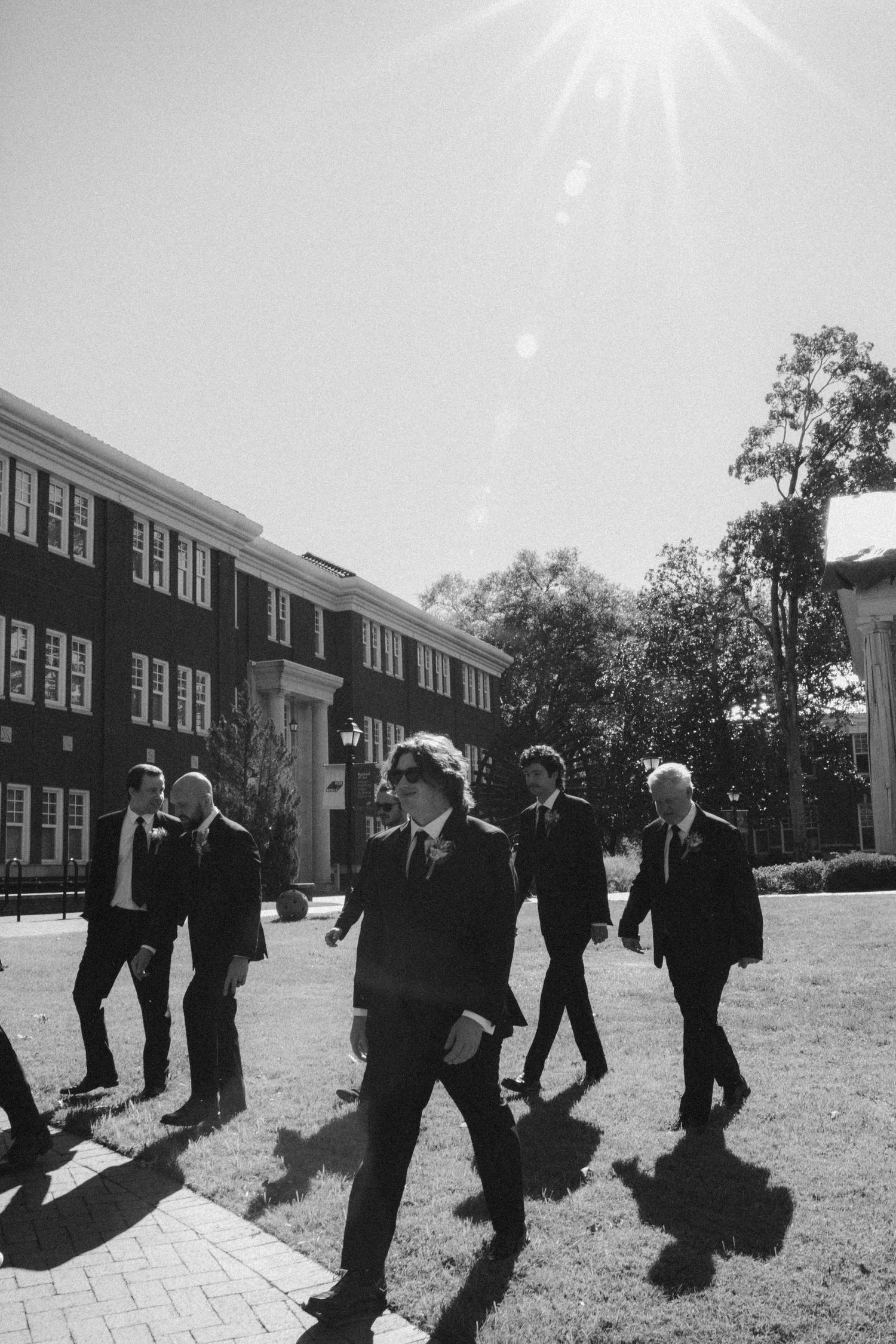 Groomsmen in formal attire walking in formation outside a brick building.