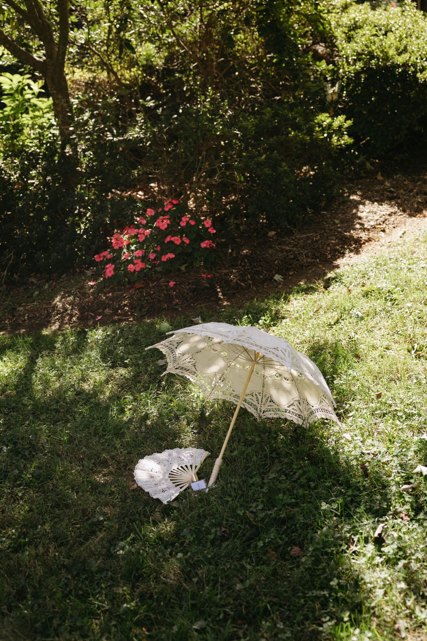 A delicate white lace parasol rests on grass near pink flowers in a sunlit garden setting.