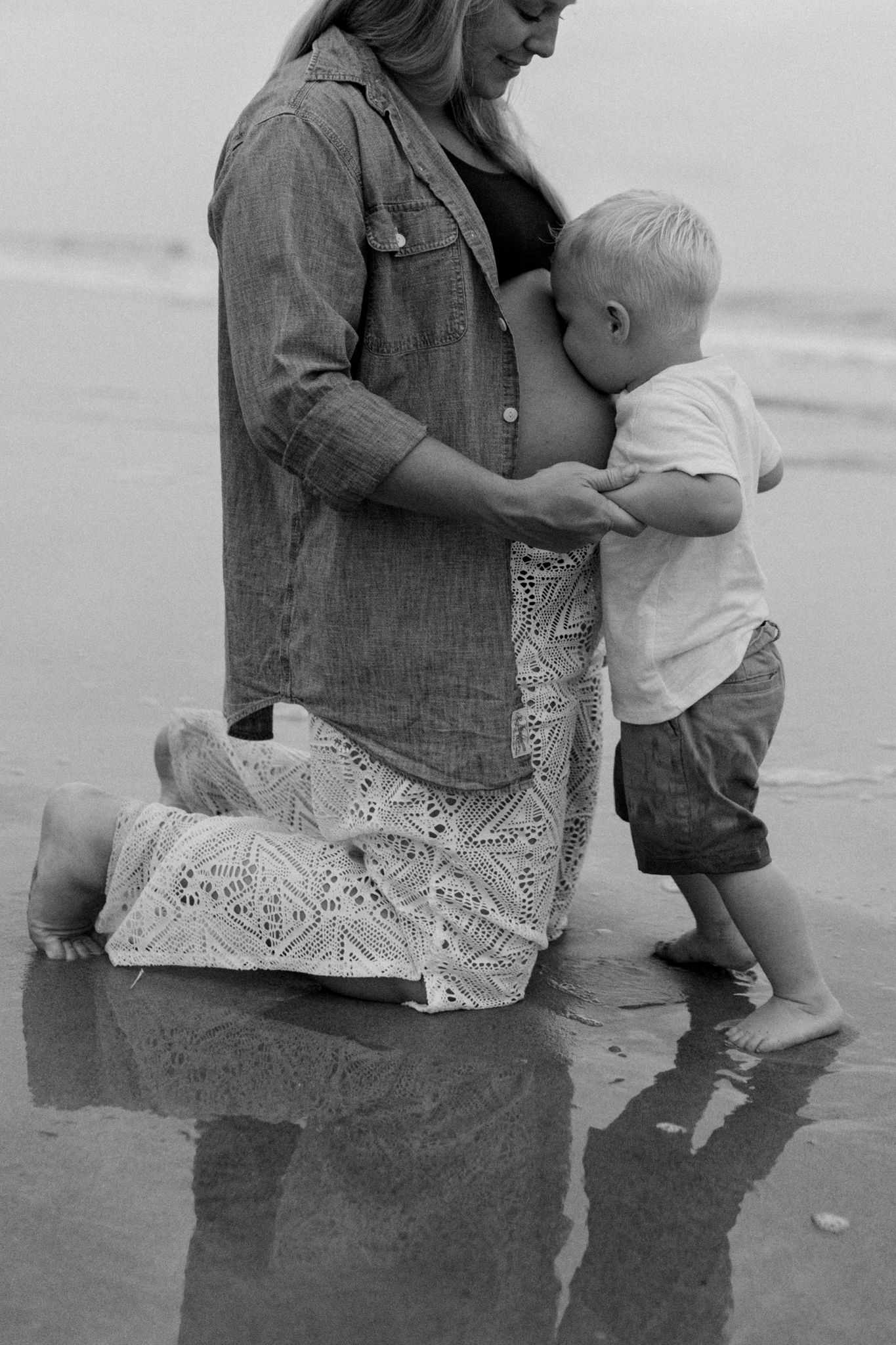 Black and white image of parent and child sitting by water's edge in vintage clothing.